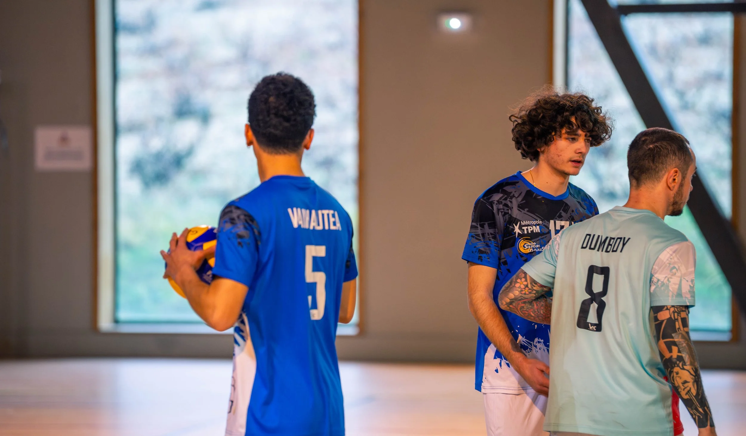 Trois joueurs de volley-ball dans un gymnase, deux avec des maillots bleus et un avec un maillot gris, discutant et préparant le jeu.