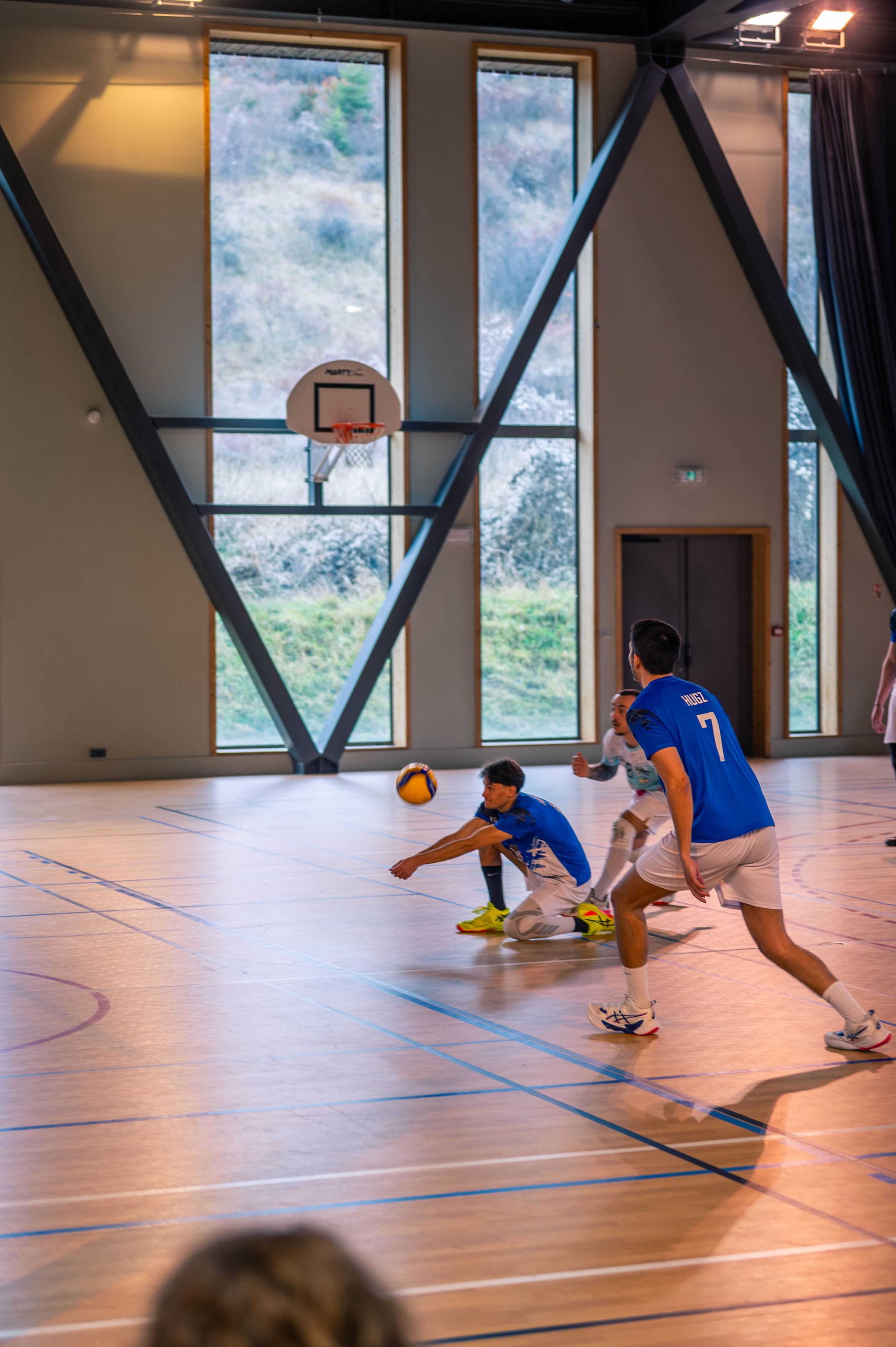 Des joueurs de volleyball en salle, un d'entre eux est sur le sol essayant de défendre, alors qu'un autre se tient prêt à jouer, dans un gymnase avec des grandes fenêtres et un panier de basketball suspendu au plafond.