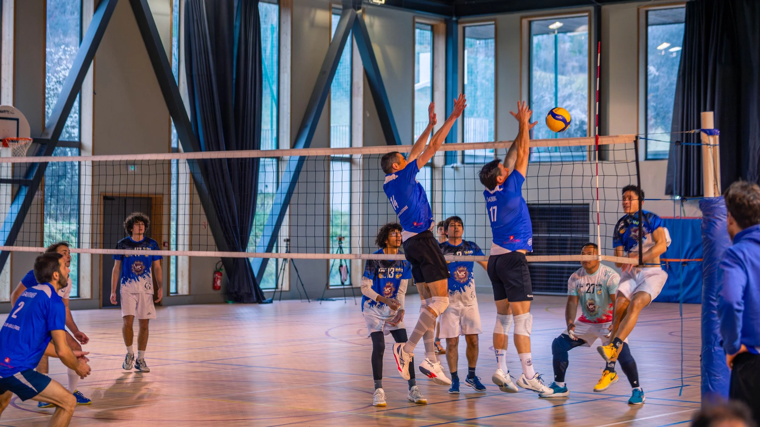 Une équipe de volley-ball joue à l'intérieur d'un gymnase. Deux joueurs en bleu sautent pour bloquer la balle, tandis qu'un joueur en blanc tente de la frapper. D'autres joueurs observent ou se préparent, avec un arbitre à droite.