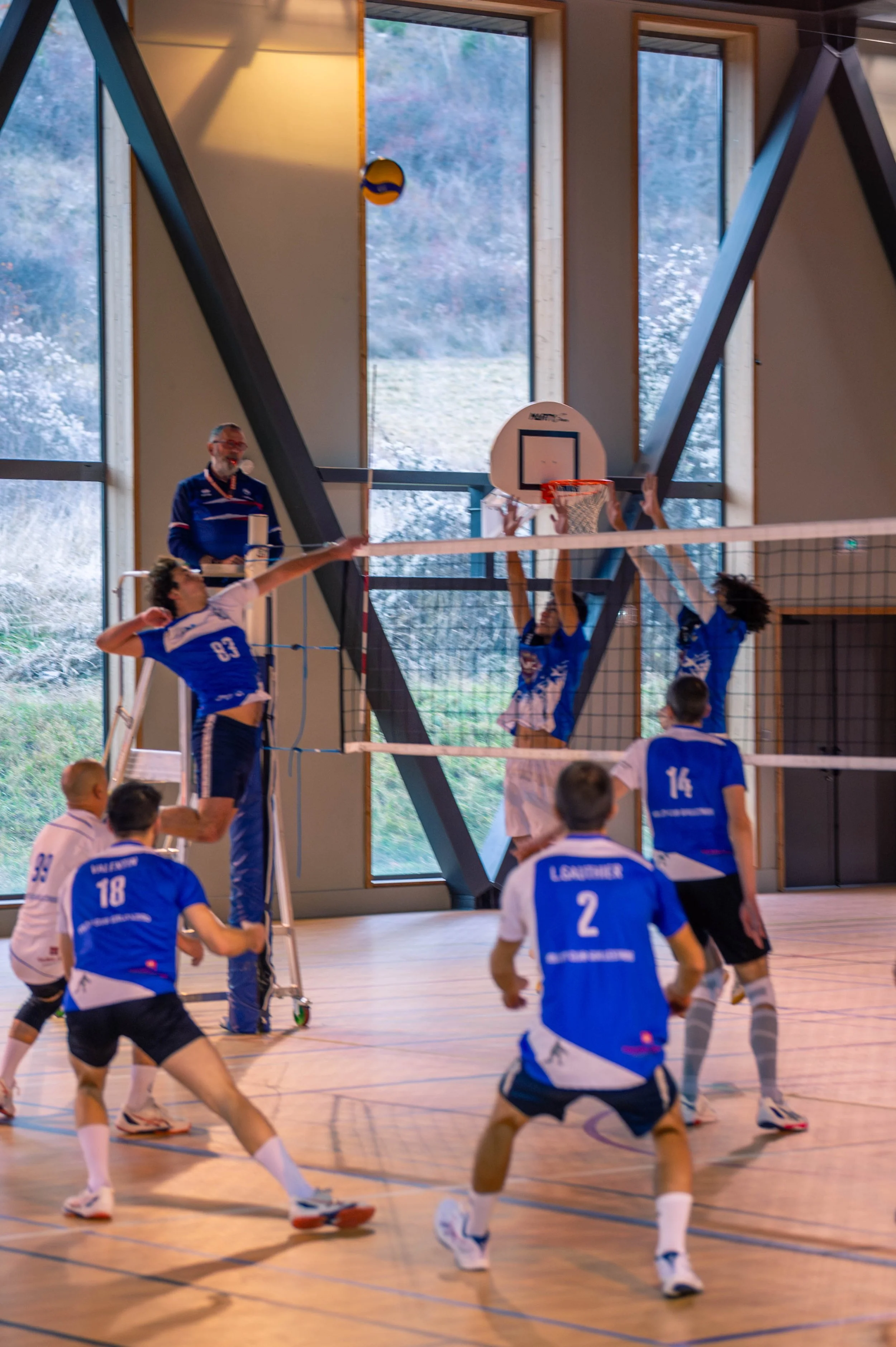 Un match de volleyball en intérieur, avec des joueurs en action, un arbitre et un public en arrière-plan.