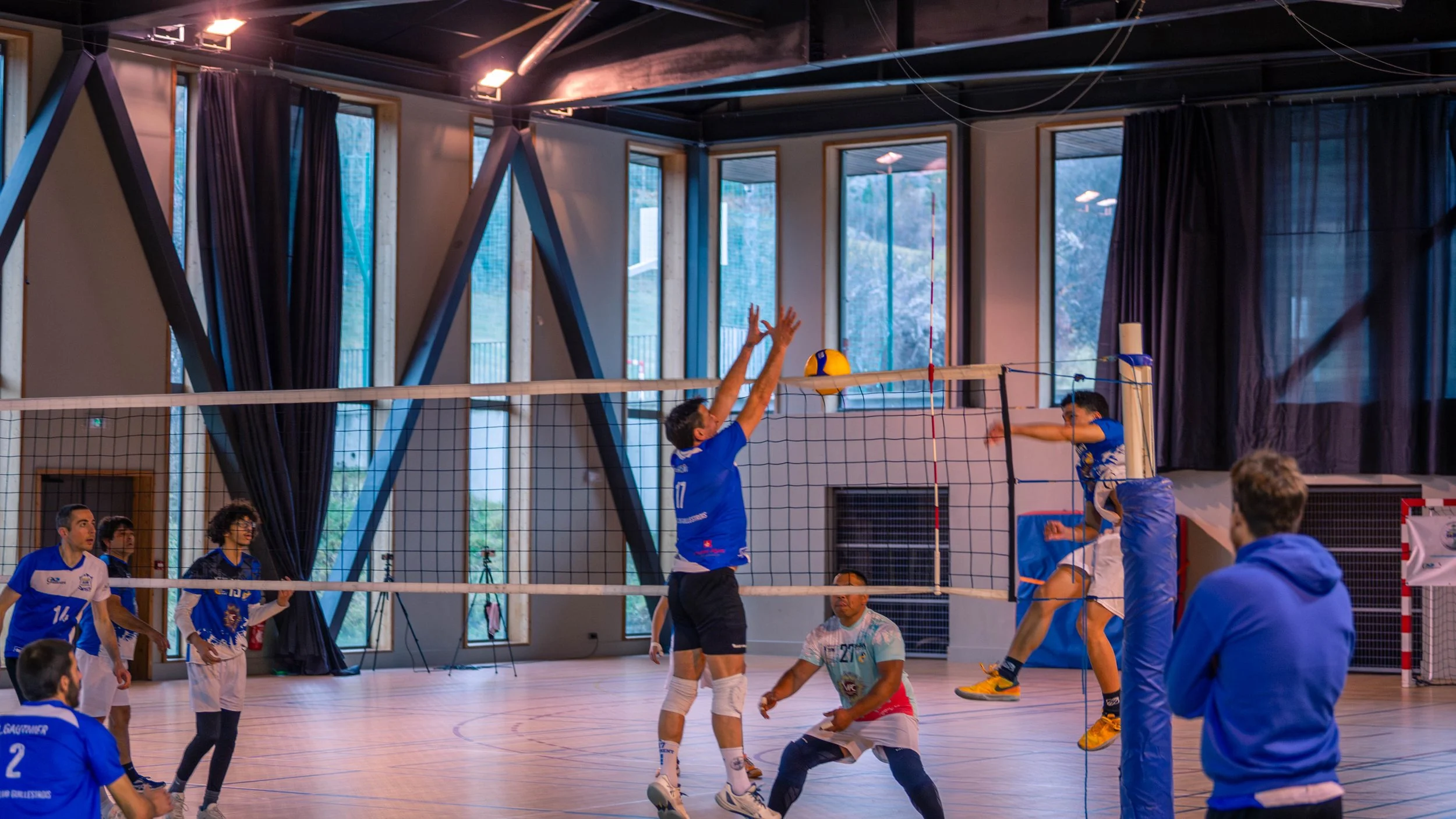 Une équipe de volleyball en pleine action dans un gymnase intérieur, avec un joueur en saut pour tenter de marquer et un joueur adverse en position de blocage.
