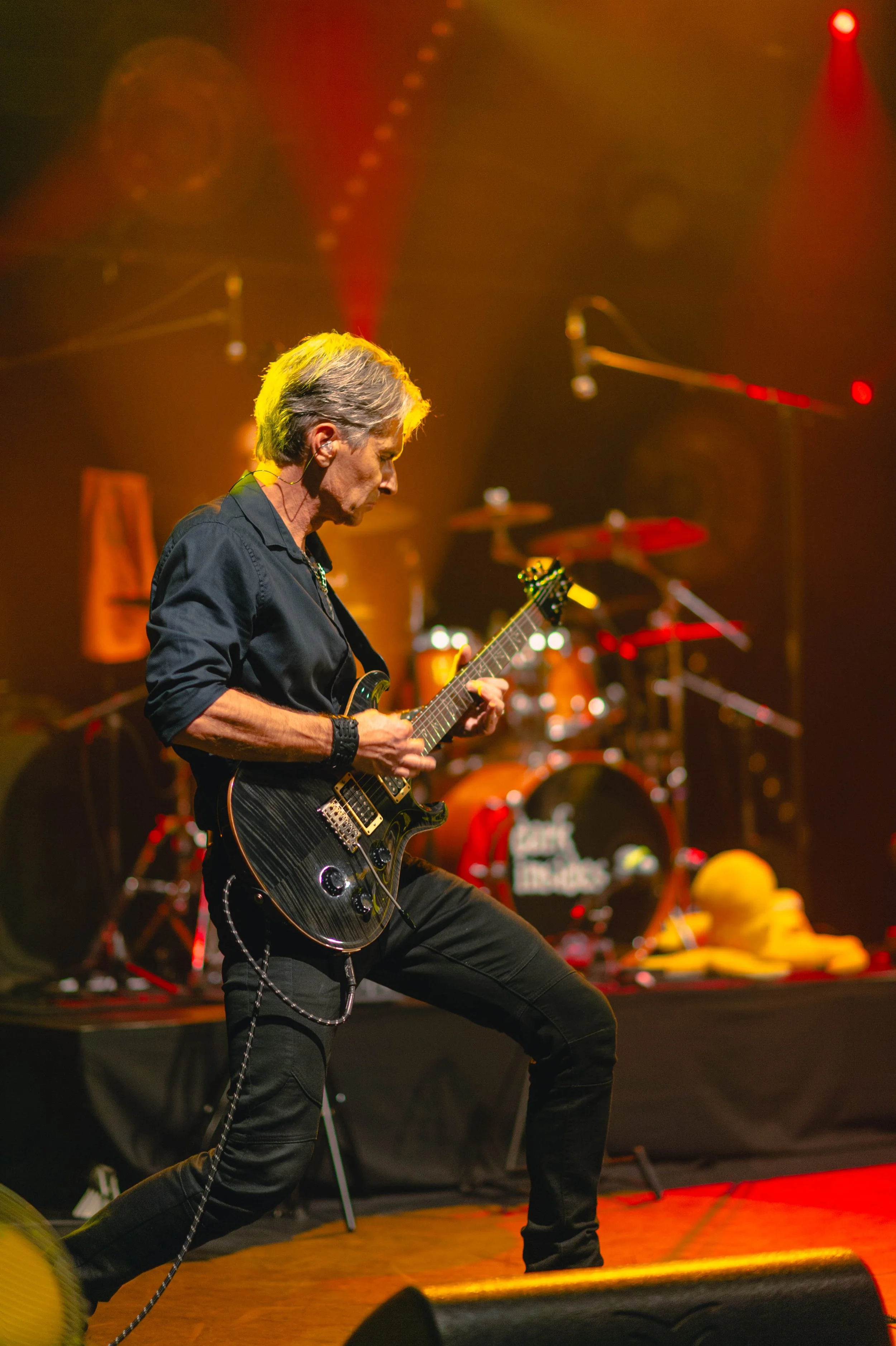 Un guitariste sur scène lors d'un concert avec un fond d'éclairage rouge et orange, un set de batterie en arrière-plan et un objet jaune au sol.