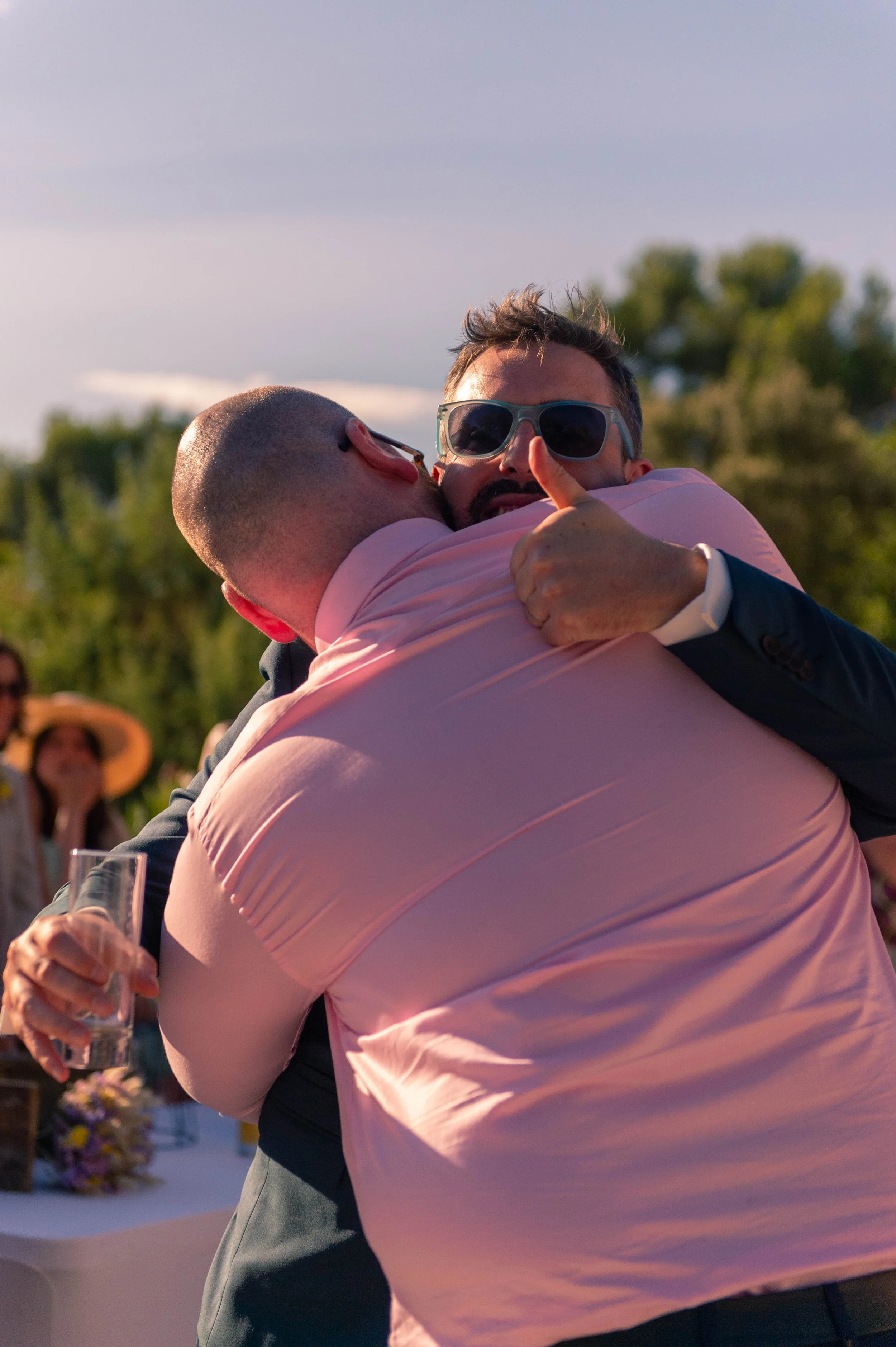 Deux hommes en costume s'embrassent chaleureusement lors d'une célébration en plein air, l'un d'eux portant des lunettes de soleil.