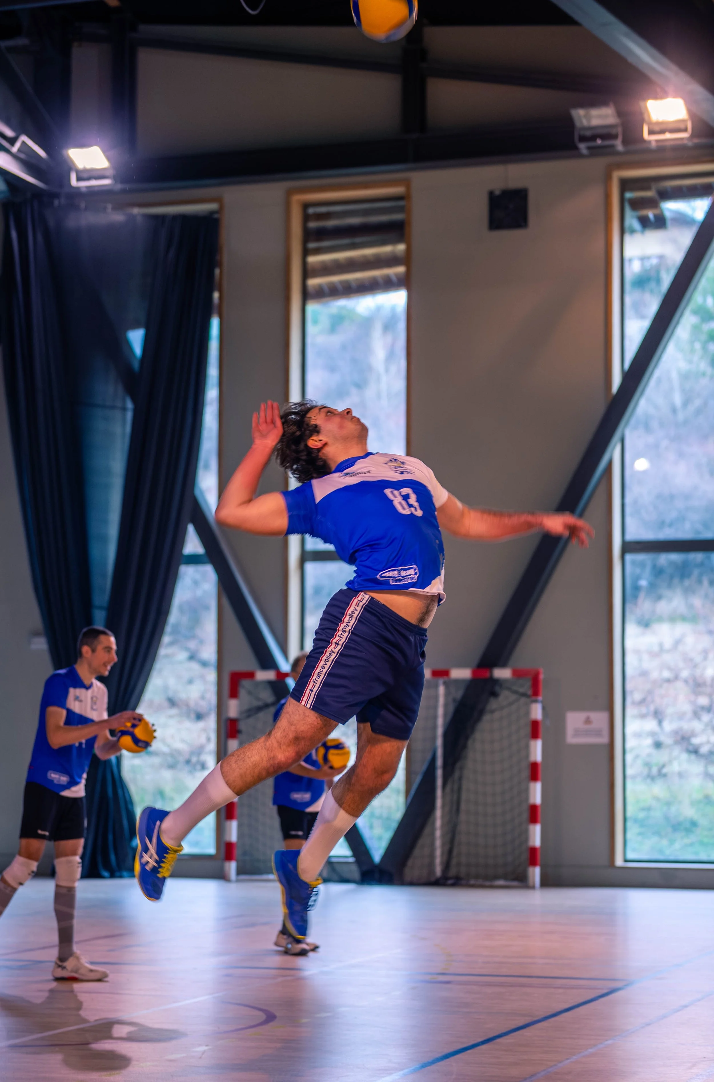 Un joueur de volleyball en pleine détente dans un gymnase, sautant pour frapper le ballon, avec d'autres joueurs en arrière-plan.