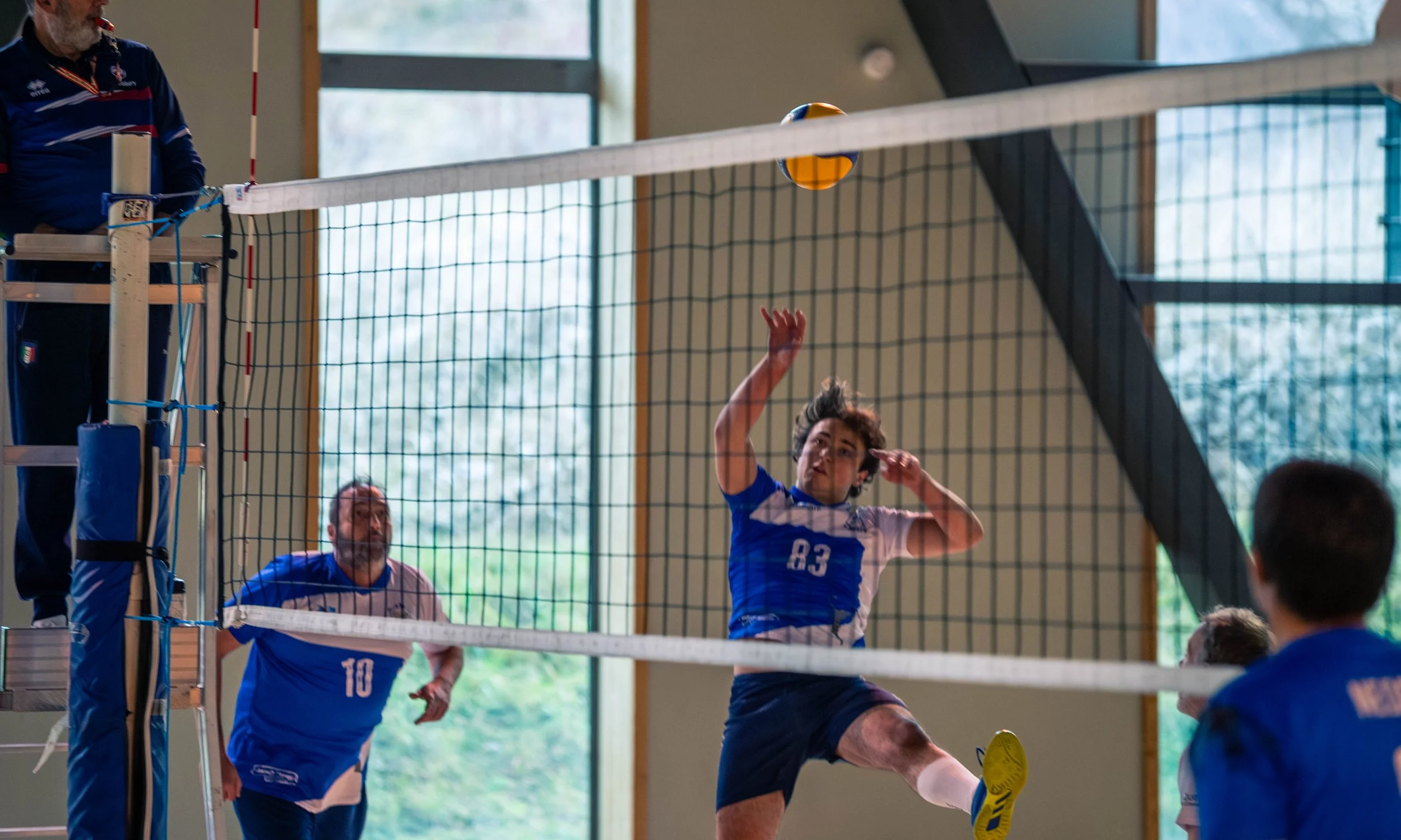 Joueur de volley-ball en action sautant pour frapper le ballon au filet lors d'un match en intérieur, autres joueurs en arrière-plan, spectateurs observant.