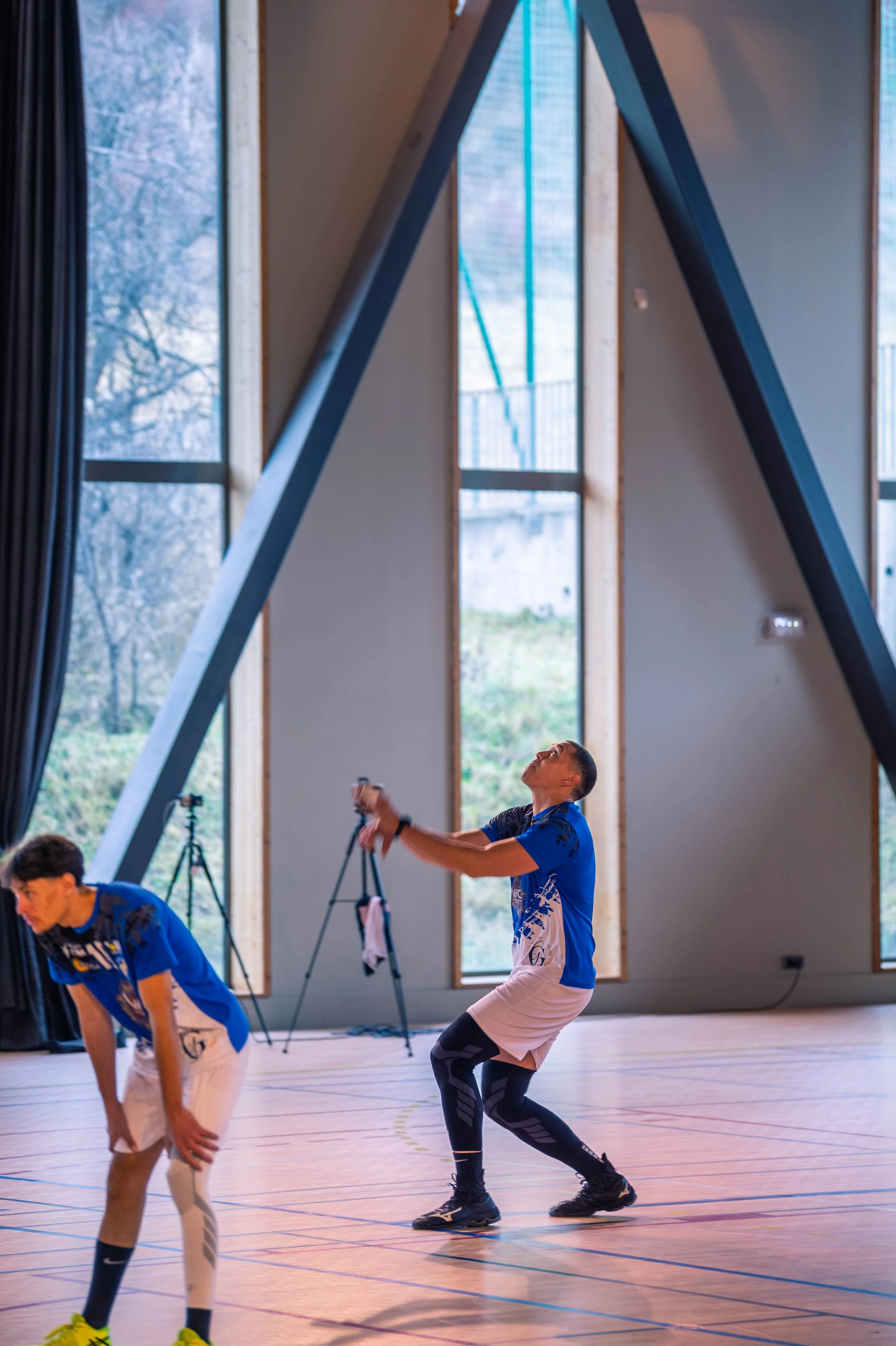 Deux joueurs de volleyball masculins dans une salle de gym, l'un en position de réception, l'autre prêt à recevoir ou à faire un sauté, avec grandes fenêtres donnant sur l'extérieur. 
