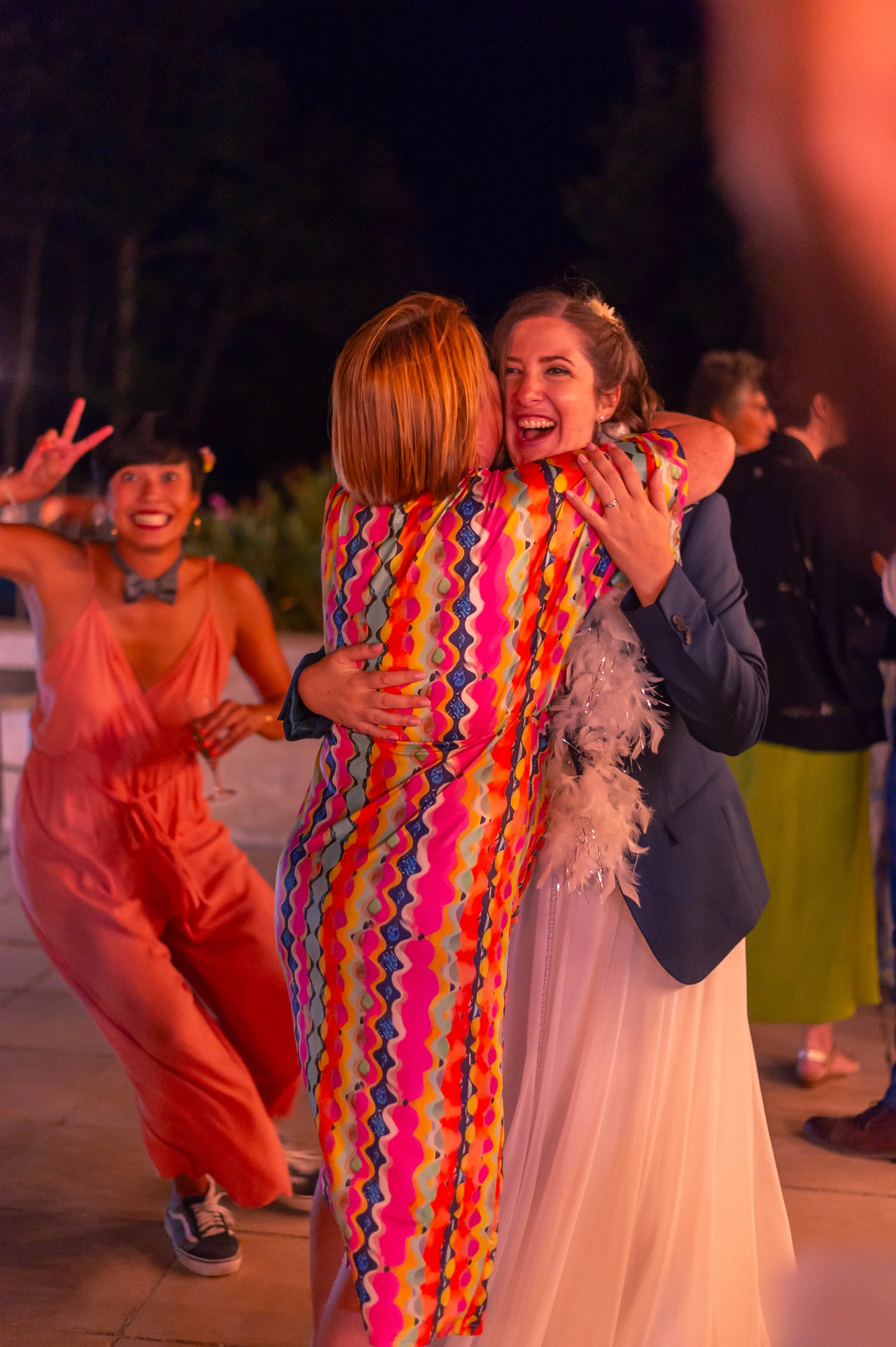 Deux femmes qui s'embrassent avec des sourires lors d'une fête en plein air la nuit. En arrière-plan, une autre femme danse et fait un signe de paix.