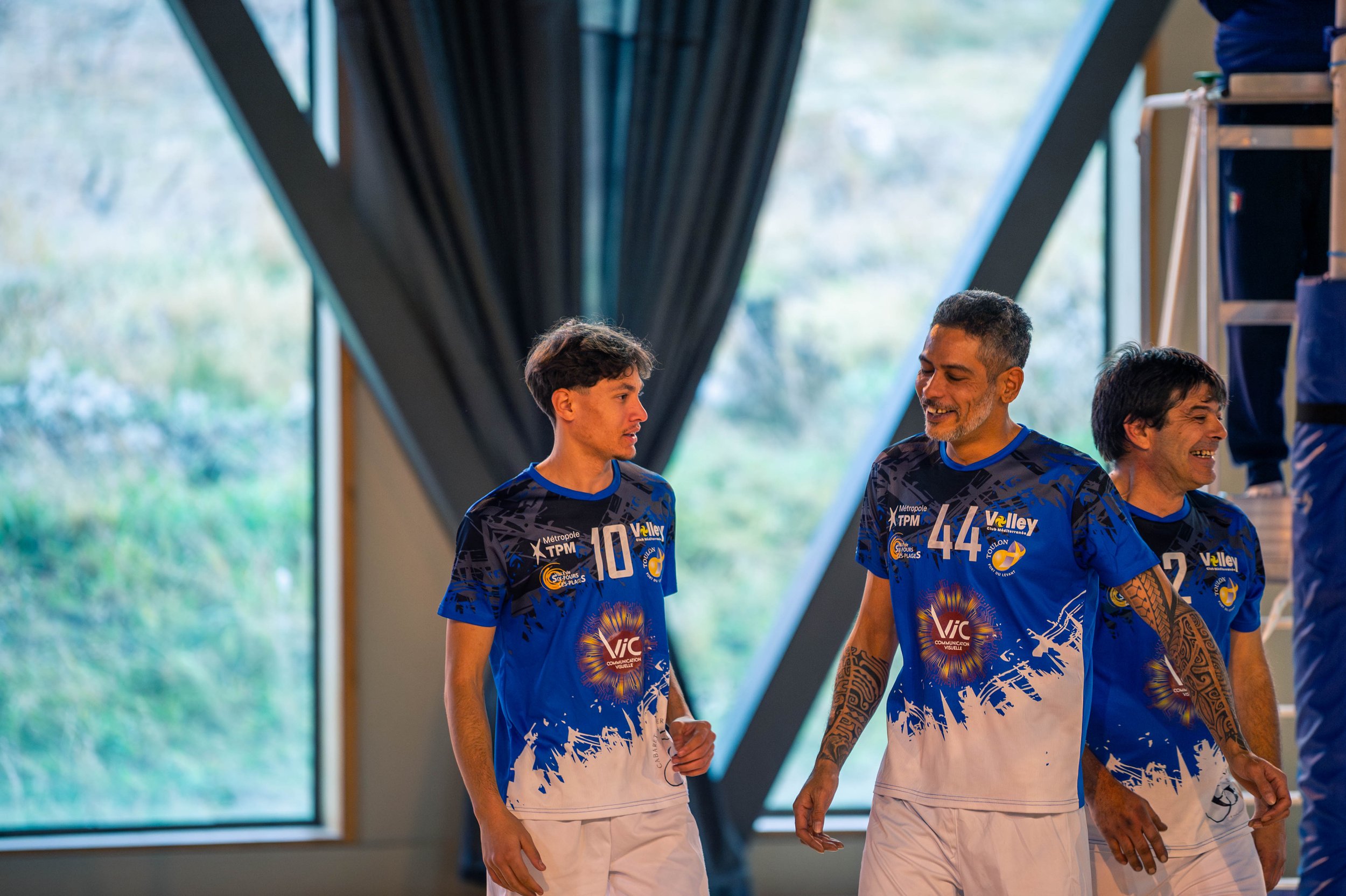 Trois joueurs de volley-ball souriants en tenue bleue avec des motifs blancs et des numéros 10, 44 et 2, dans une salle d'entraînement près de grandes fenêtres extérieures.