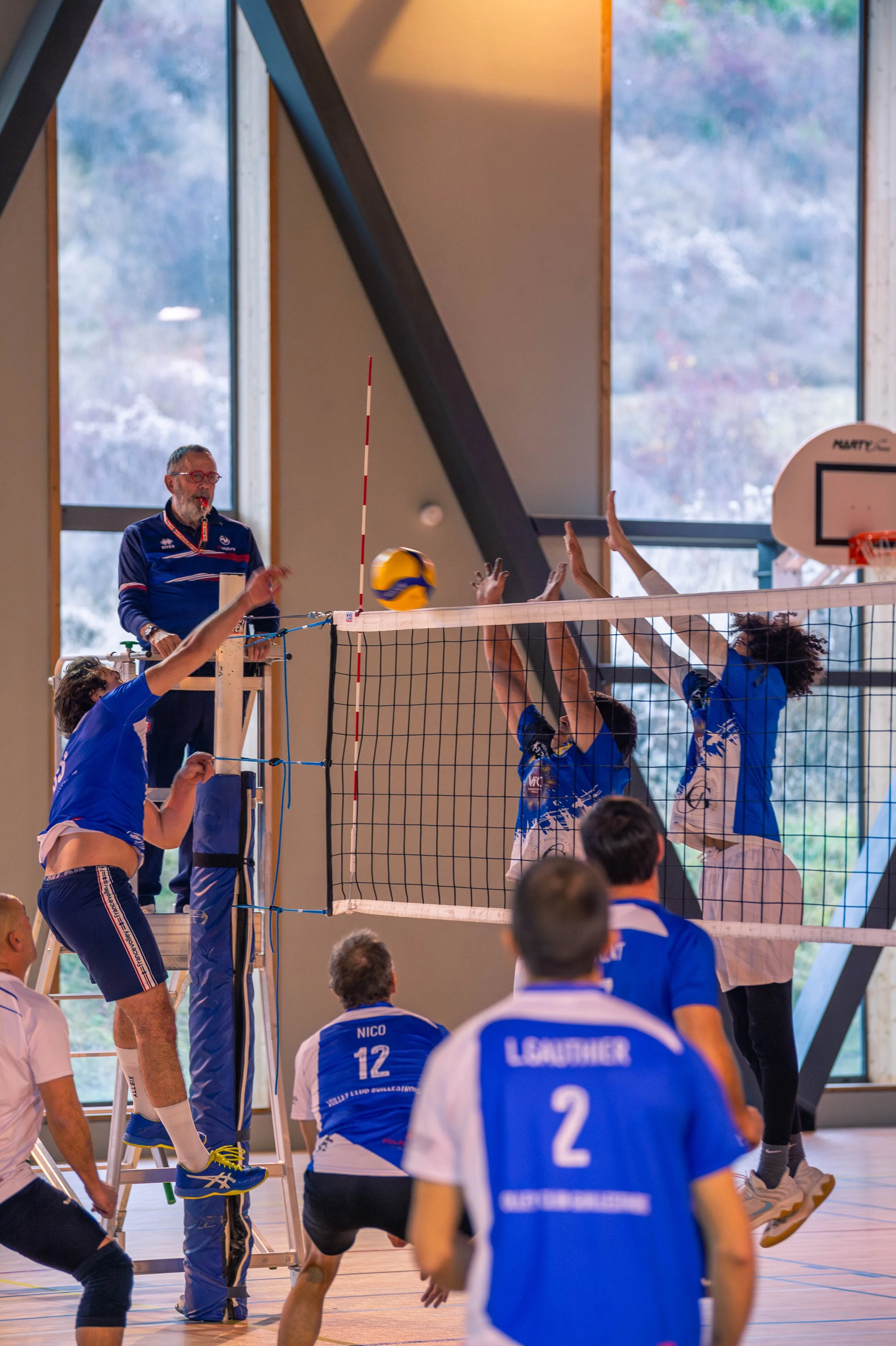 Une photo d'un match de volley-ball en intérieur, avec des joueurs en tenues bleues. Un arbitre se tient sur une chaise et observe le jeu, tandis que joueurs tentent de bloquer ou de frapper le ballon près du filet.