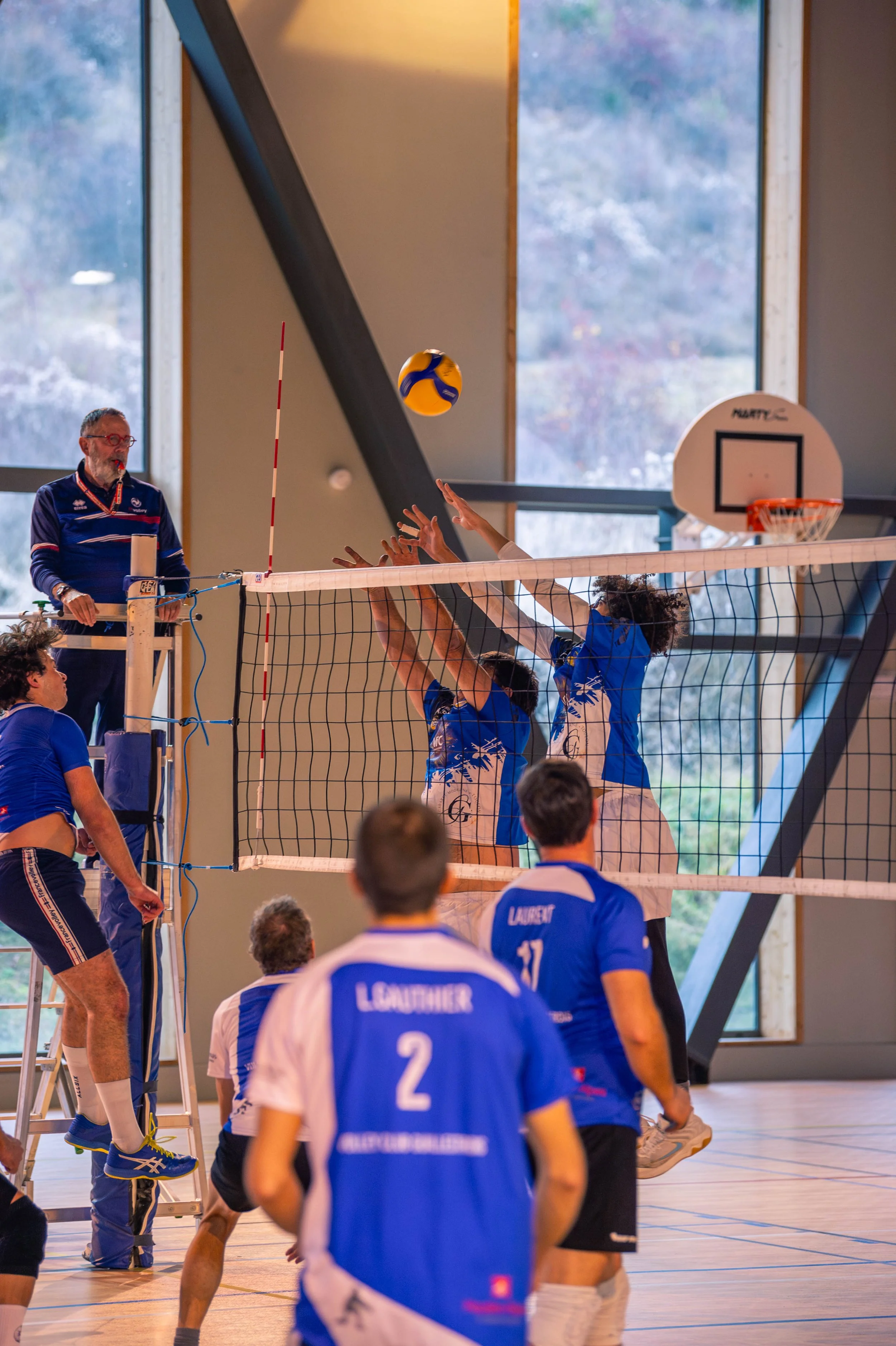 Joueurs de volley-ball en action lors d'un match intérieur, un joueur au premier plan porte un maillot bleu avec le nom 'LAURET' et le numéro 2. Deux joueurs au-dessus tapent le ballon en haut du filet, un arbitre se tient à côté.