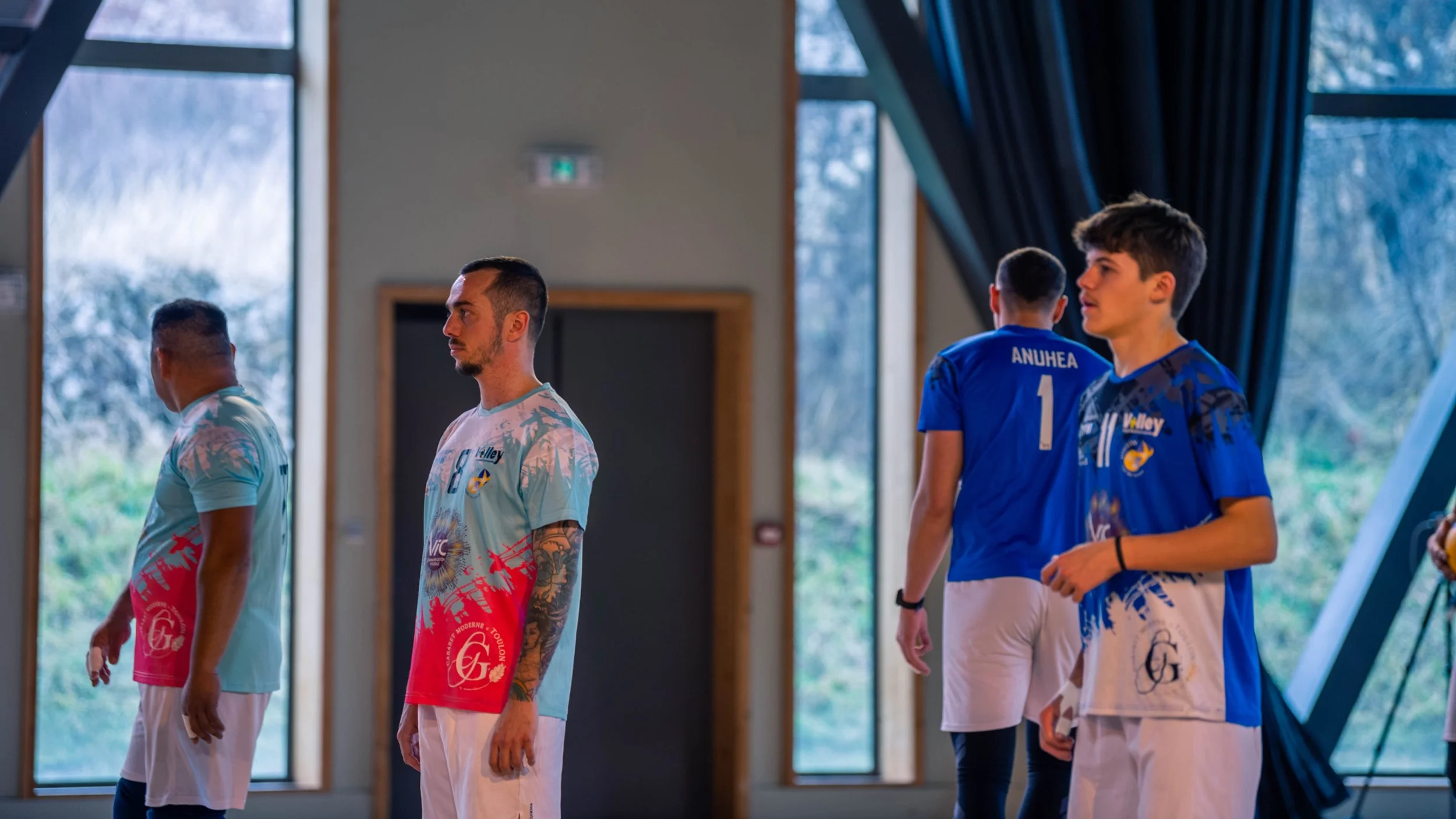 Jeunes sportifs dans une salle, portant des maillots colorés, certains ont des tatouages visibles sur les bras, en arrière-plan des fenêtres laissant entrevoir un paysage naturel.