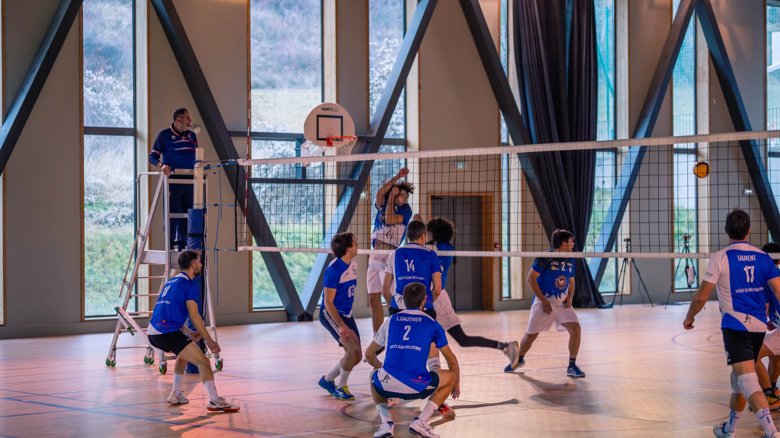 Une équipe de volley-ball en train de jouer dans une salle intérieure, avec un joueur sautant pour frapper le ballon au-dessus du filet, tandis que d'autres joueurs se préparent en position défensive.