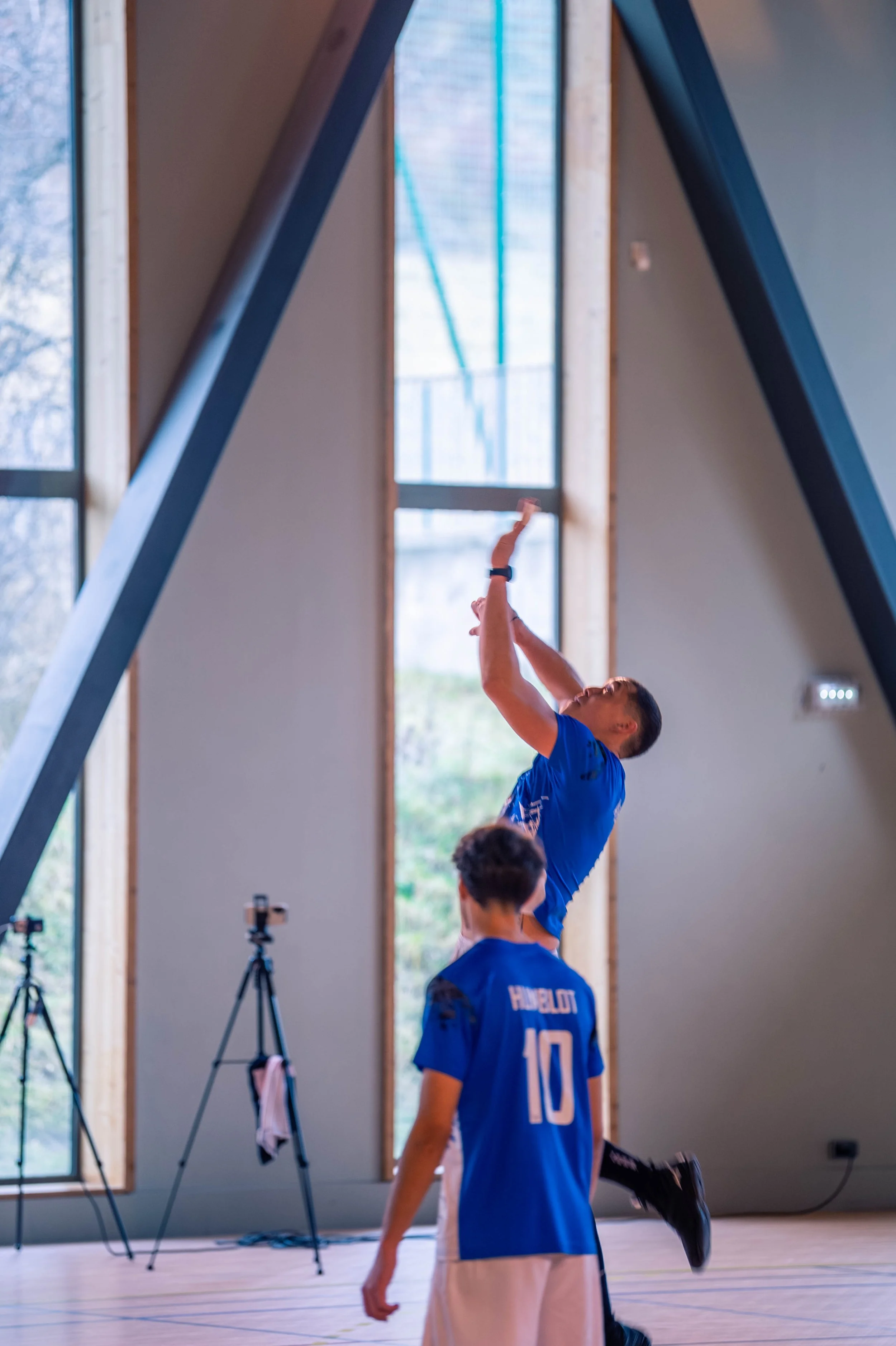 Deux jeunes hommes en maillots de sport bleus, un sautant en l'air et l'autre marchant, dans une salle de sport avec de grandes fenêtres.