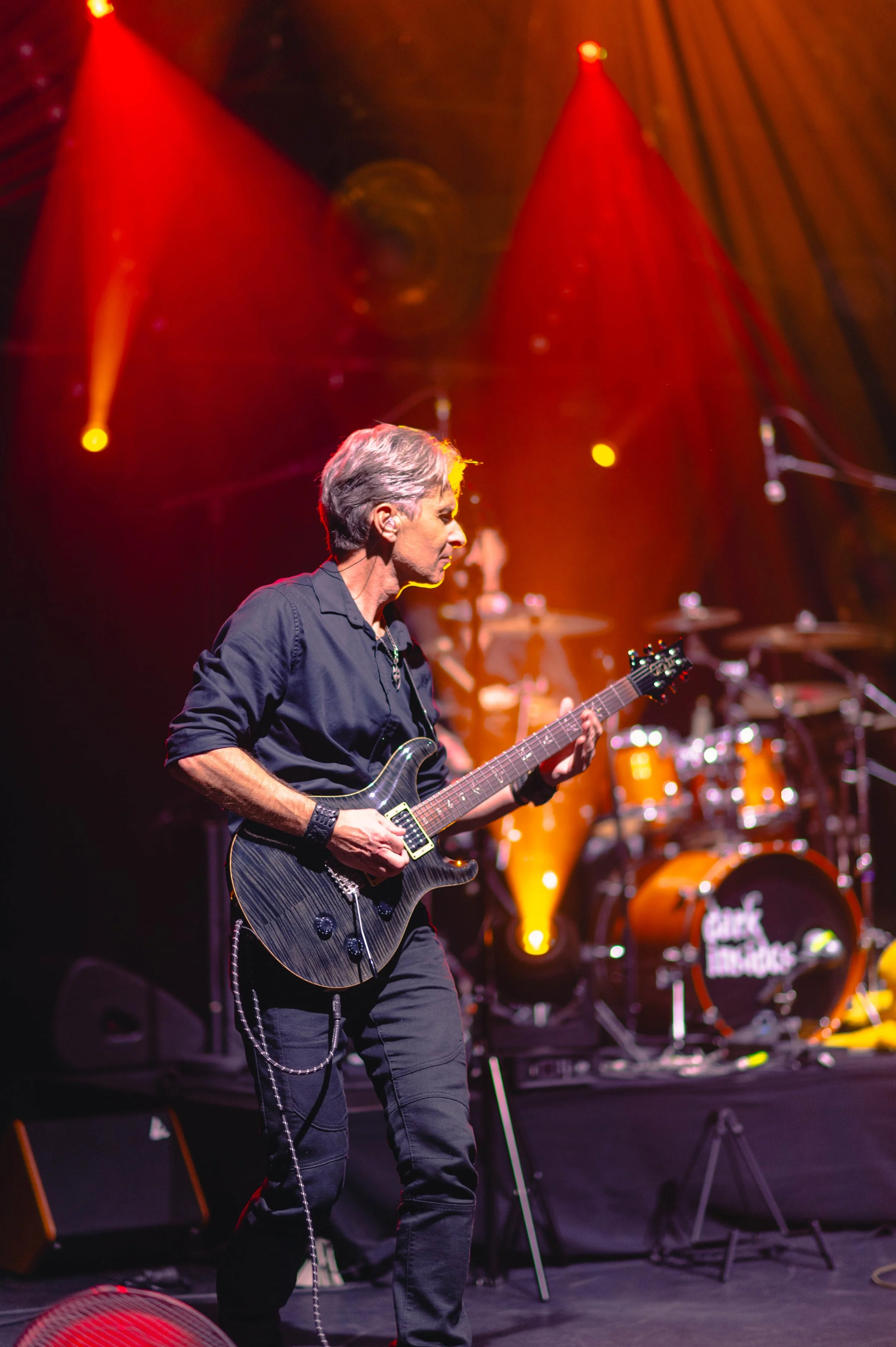 Un musicien joue de la guitare électrique lors d'un concert, avec un groupe de percussion en arrière-plan, sur scène avec un éclairage orange et rouge.
