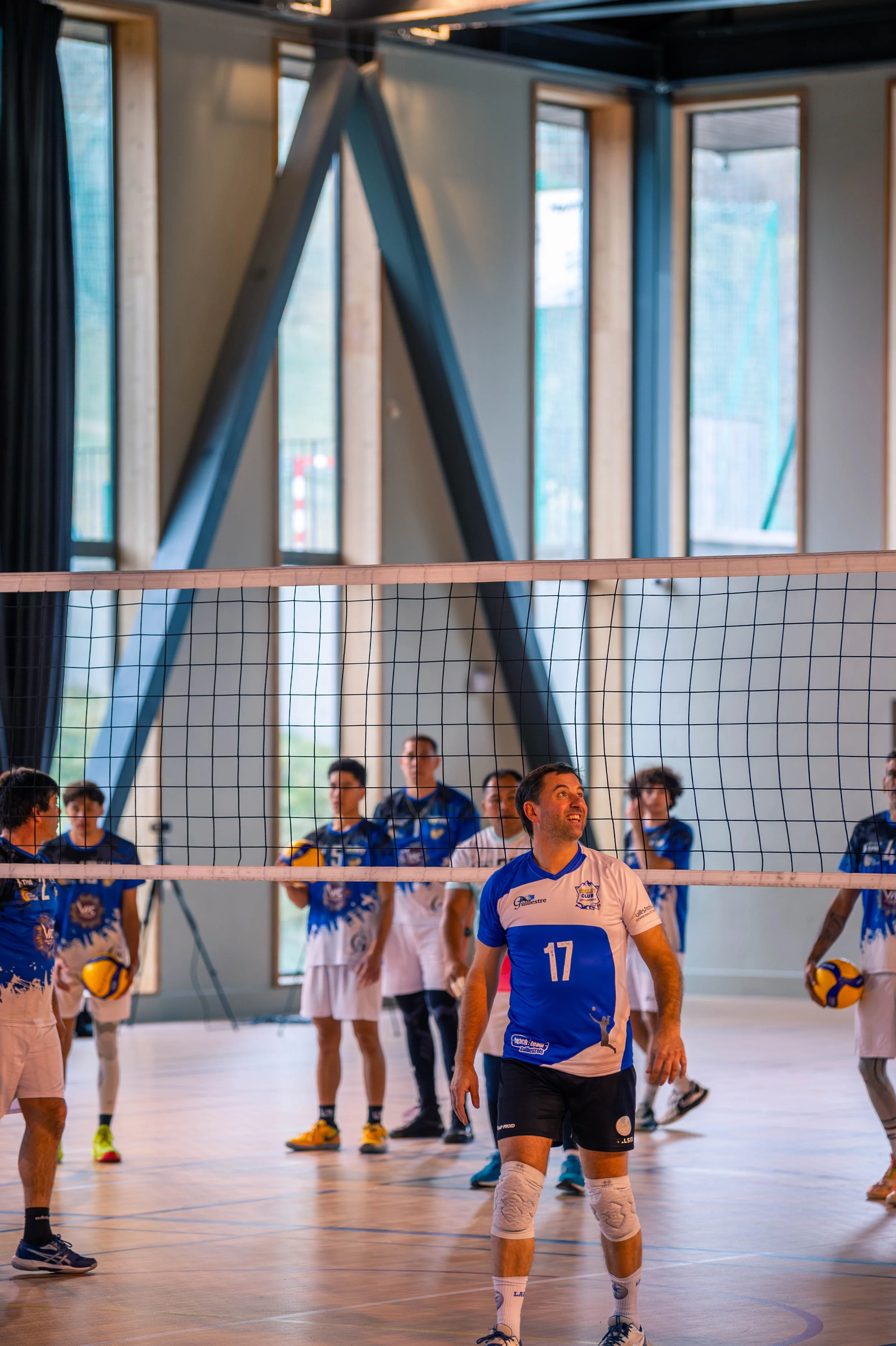 Une équipe de volleyball en entraînement dans une salle intérieure, avec un joueur en avant portant un maillot numéro 17, entouré de ses coéquipiers portant des maillots bleus et blancs, sous une structure métallique et de grandes fenêtres lumineuses