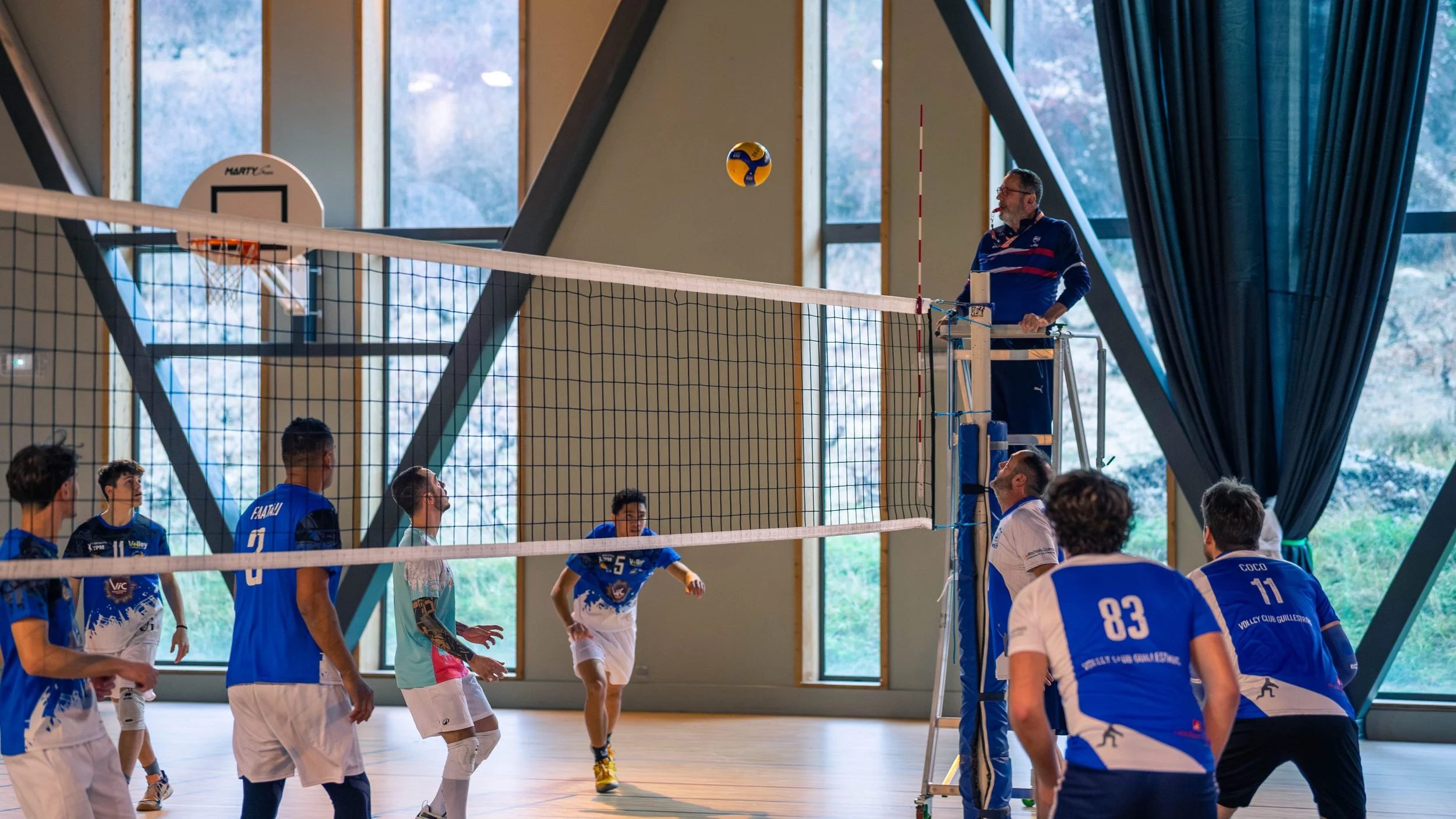 Une équipe de volley-ball en train de jouer dans une salle intérieure avec de grandes fenêtres. Un joueur effectue un service ou une frappe tandis que les autres se préparent pour recevoir ou défendre, sous la supervision d'un arbitre.