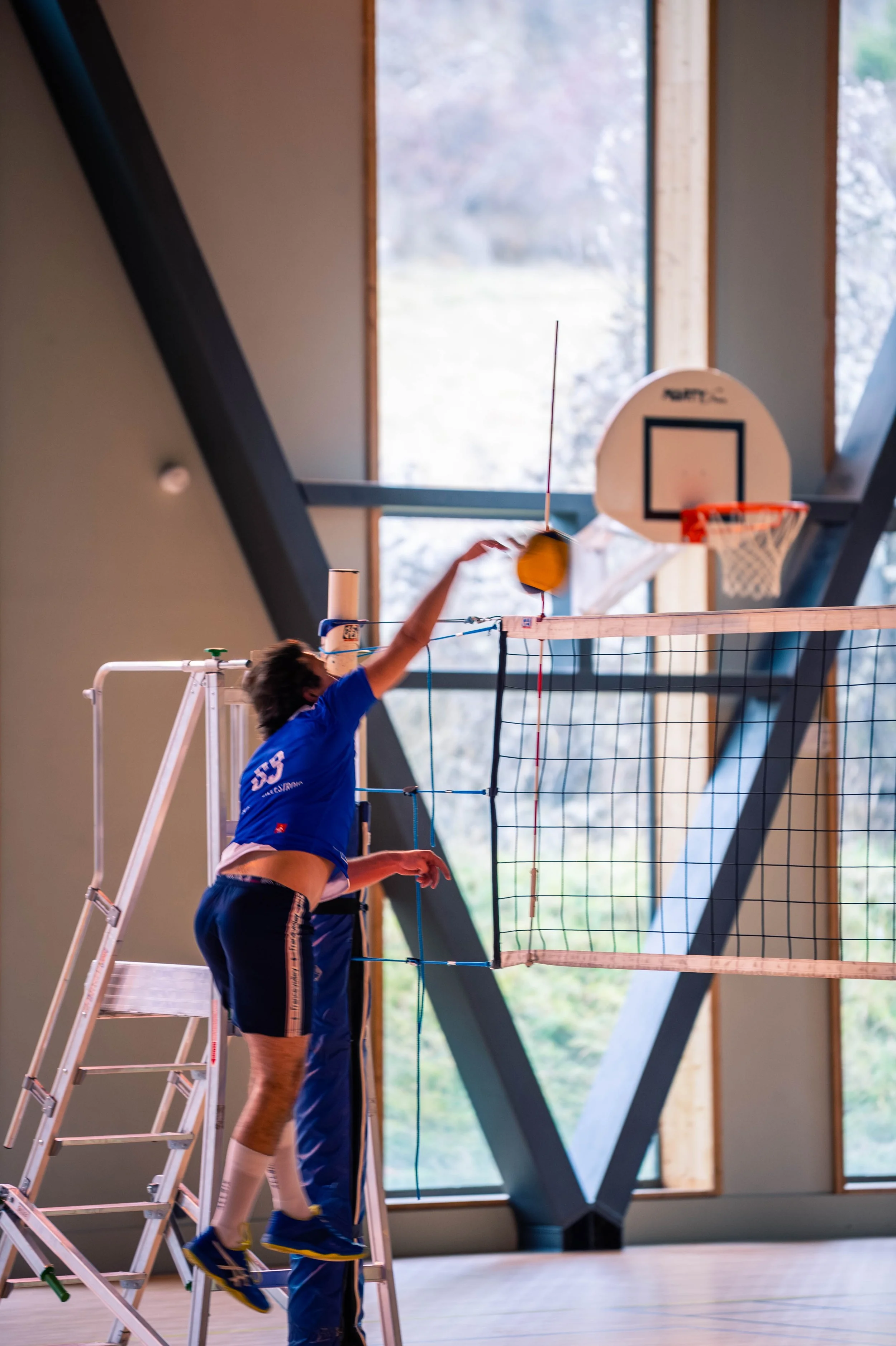 Un enfant joue au volleyball en intérieur, utilisant un filet de volleyball installé près d'une grande fenêtre dans une salle de sport.