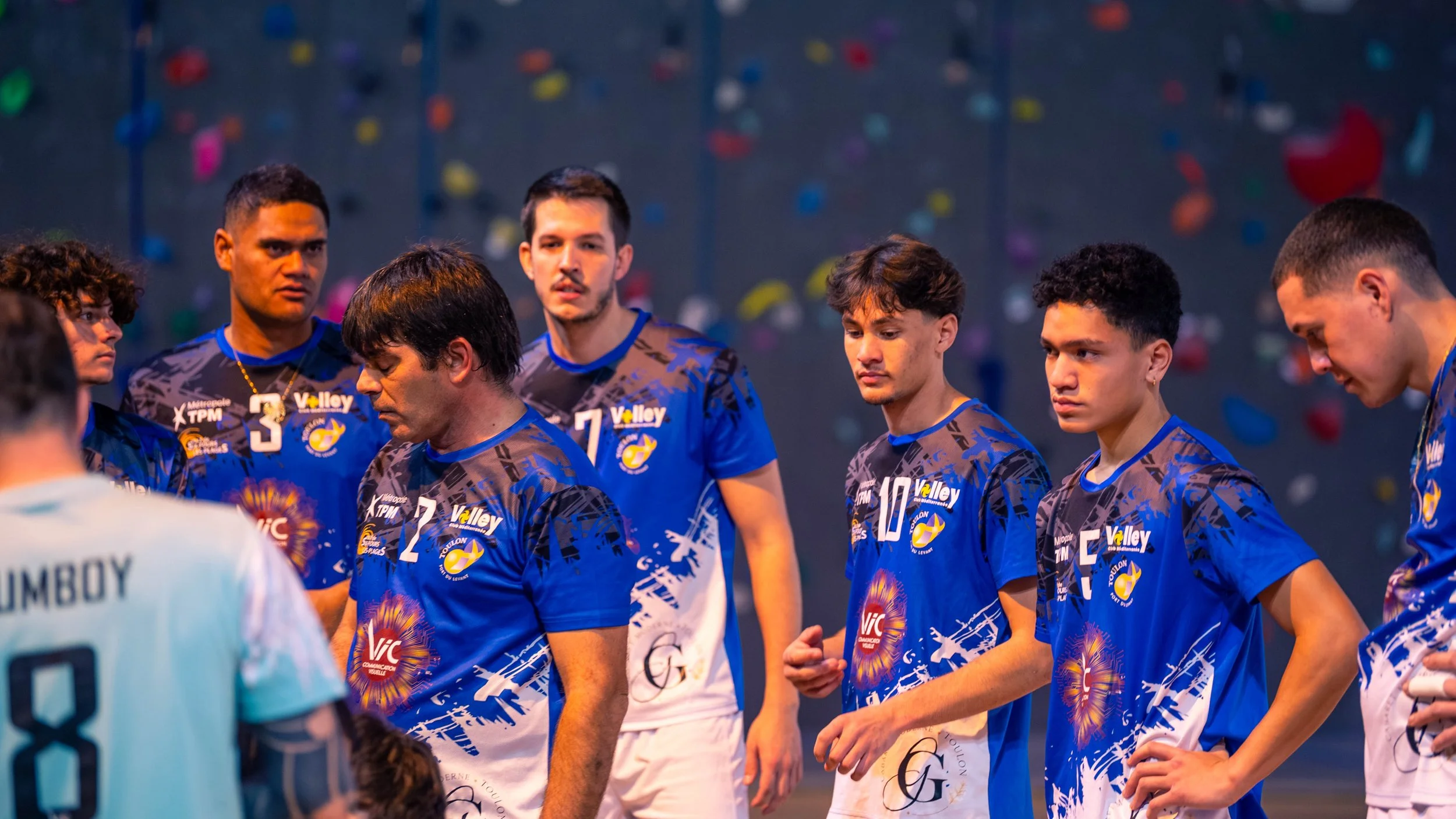 Groupe de jeunes joueurs de volleyball professionnels en tenue bleue, lors d'une séance d'équipe, sur un mur de salle d'escalade en arrière-plan.