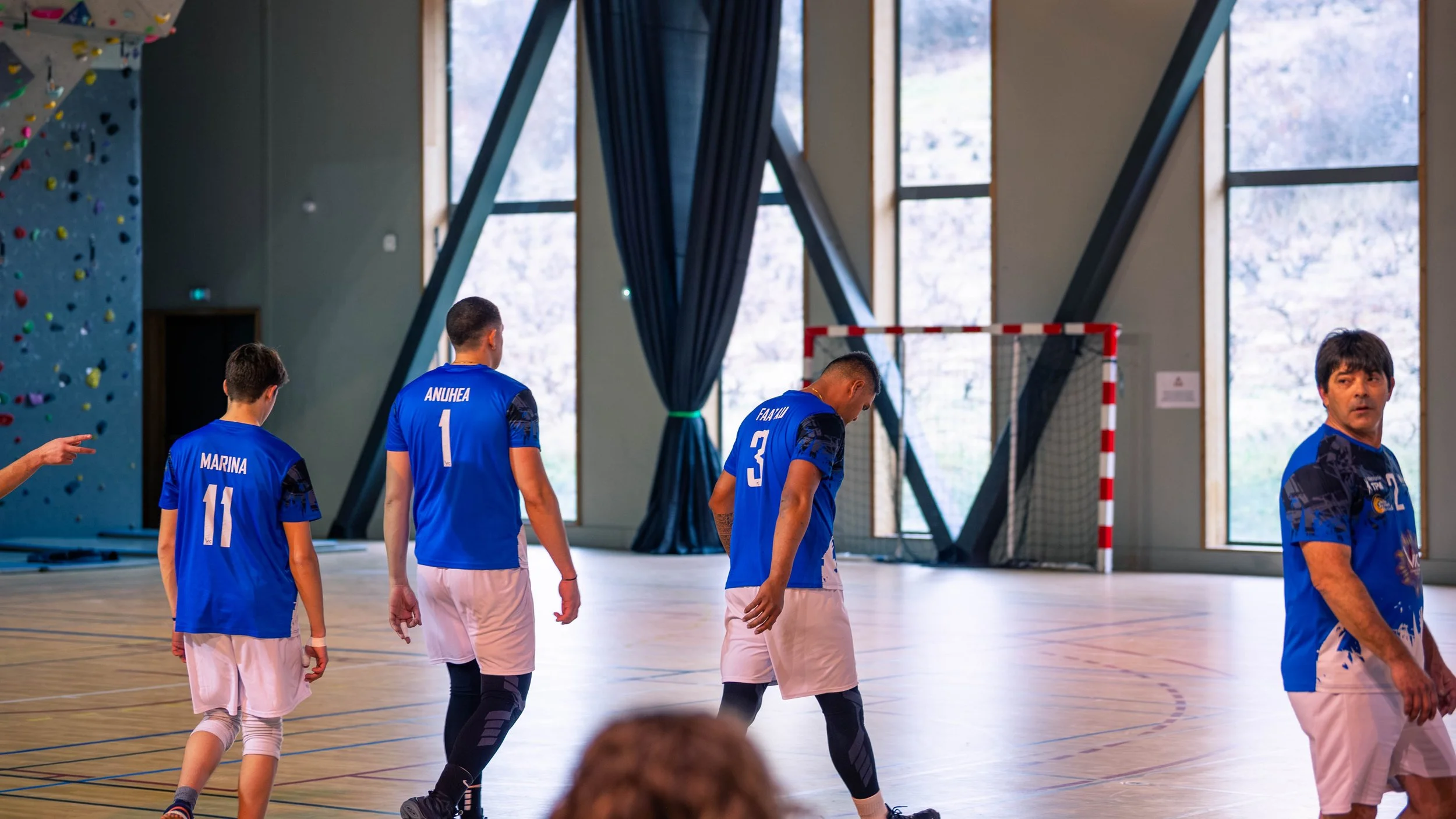 Joueur de handball dans une salle intérieure, portant un maillot bleu, marchant avec d'autres joueurs, avec un but de handball derrière eux et une paroi d'escalade à gauche.