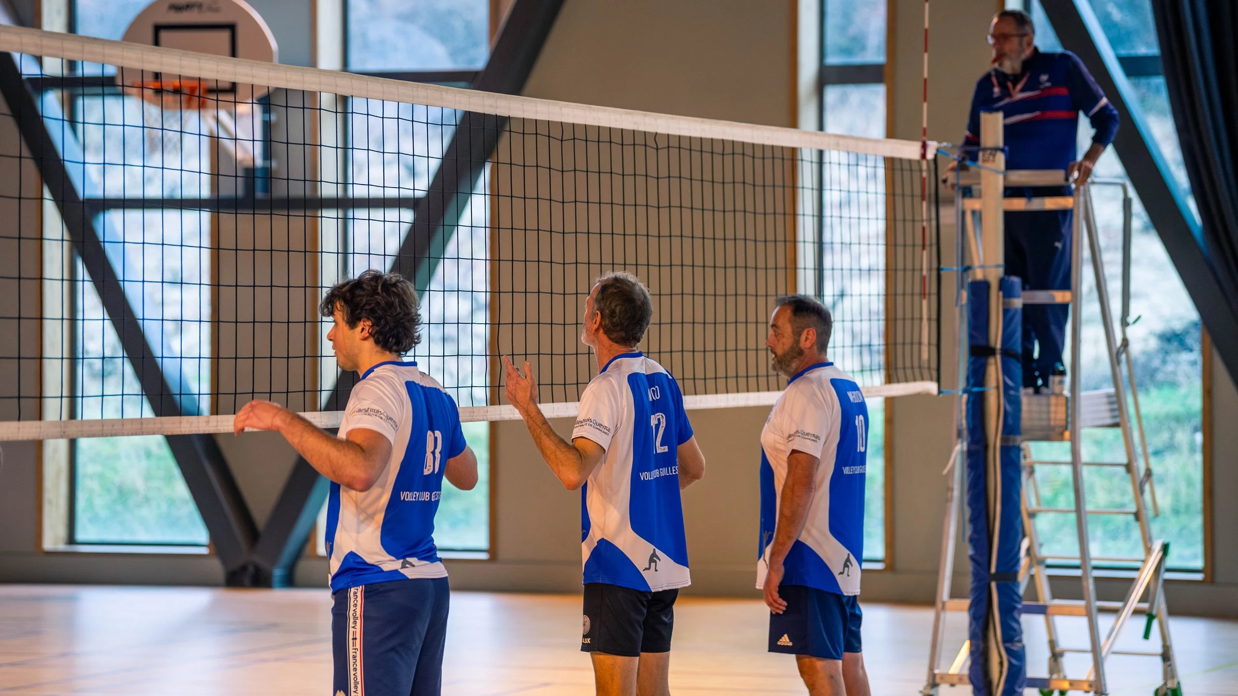 Quatre joueurs de volley-ball en tenue de sport se préparent devant un filet dans une salle de sport.