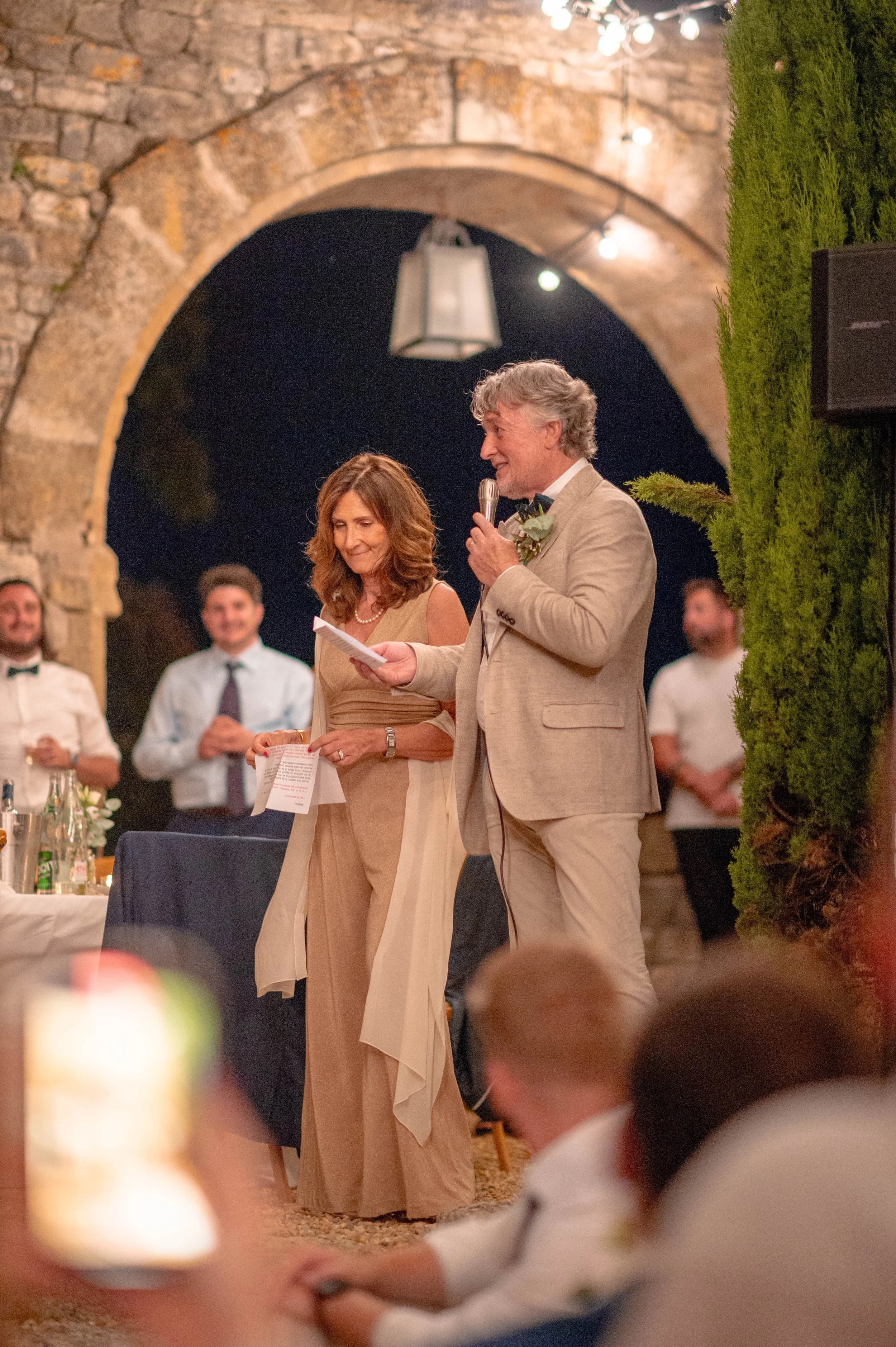 Un couple lors d’un mariage, la personne masculine lit un discours avec un microphone, autour d’eux, des invités souriants dans un décor en pierre.