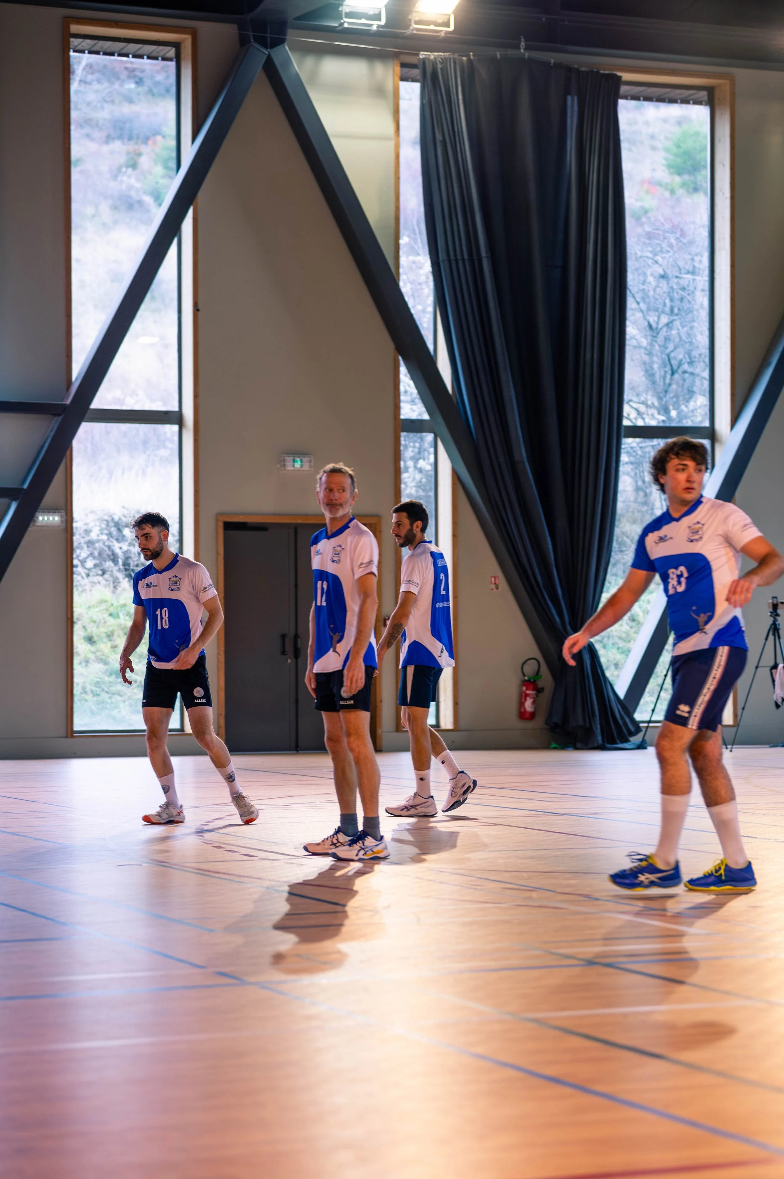 Jeu de volleyball en salle avec quatre joueurs en maillots bleus et blancs sur un parquet en bois, dans un gymnase lumineux avec de grandes fenêtres et des rideaux noirs.