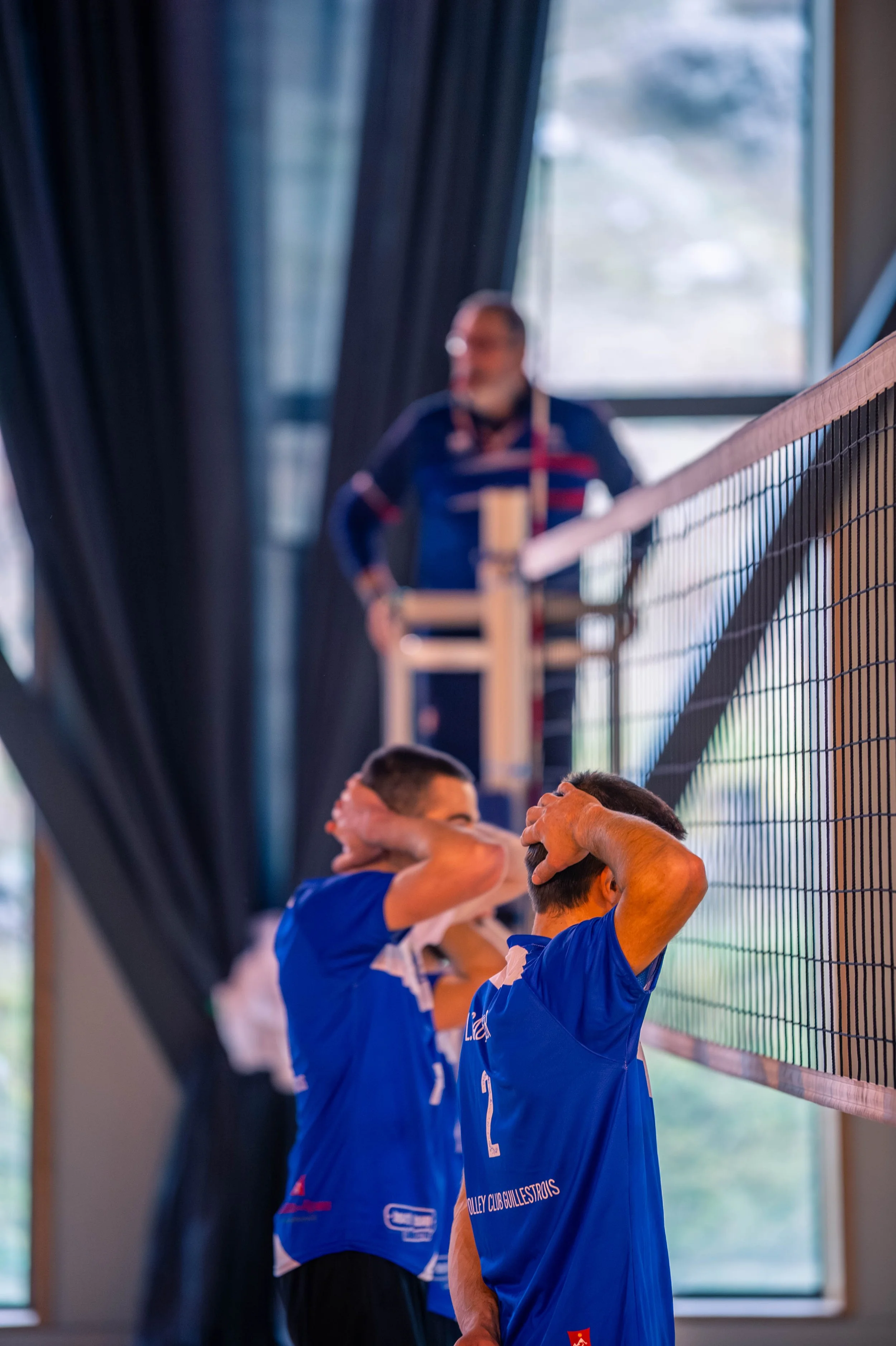 Deux joueurs de volley-ball en tenue bleue, couvrant leurs oreilles, dans une salle avec un filet et un entraîneur en arrière-plan, observant la scène