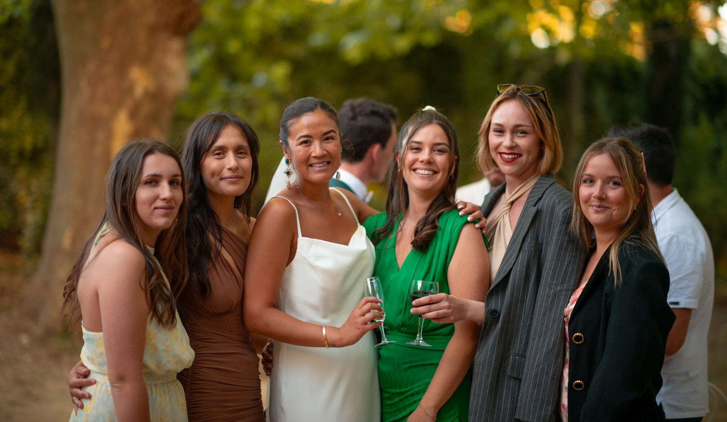 Groupe de six femmes souriantes lors d'un évènement en plein air, tenant des verres de vin rouge, avec un fond de nature et d'arbres.