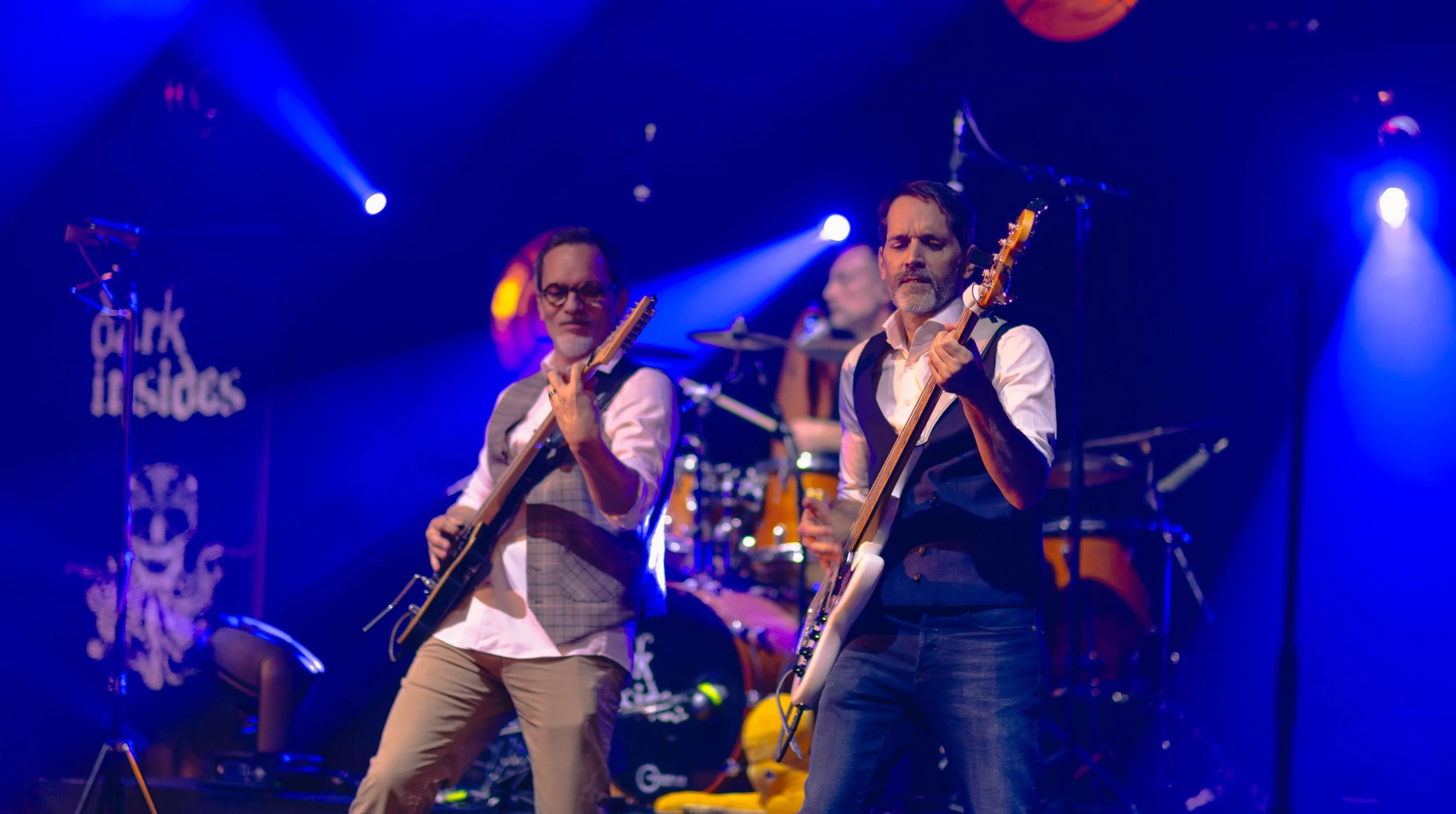 Un groupe de musiciens joue sur scène sous des lumières bleues. Deux guitaristes en avant jouent de leurs instruments, un batteur en arrière joue derrière sa batterie.