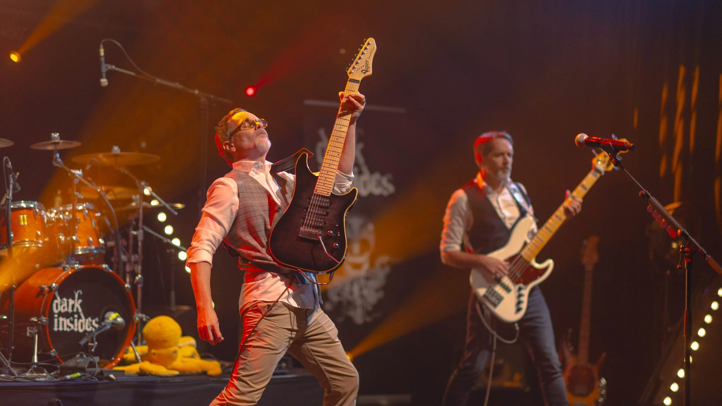 Deux musiciens jouent sur scène avec des guitares électriques, entourés d'équipements de musique, sous un éclairage de scène à couleur chaude.