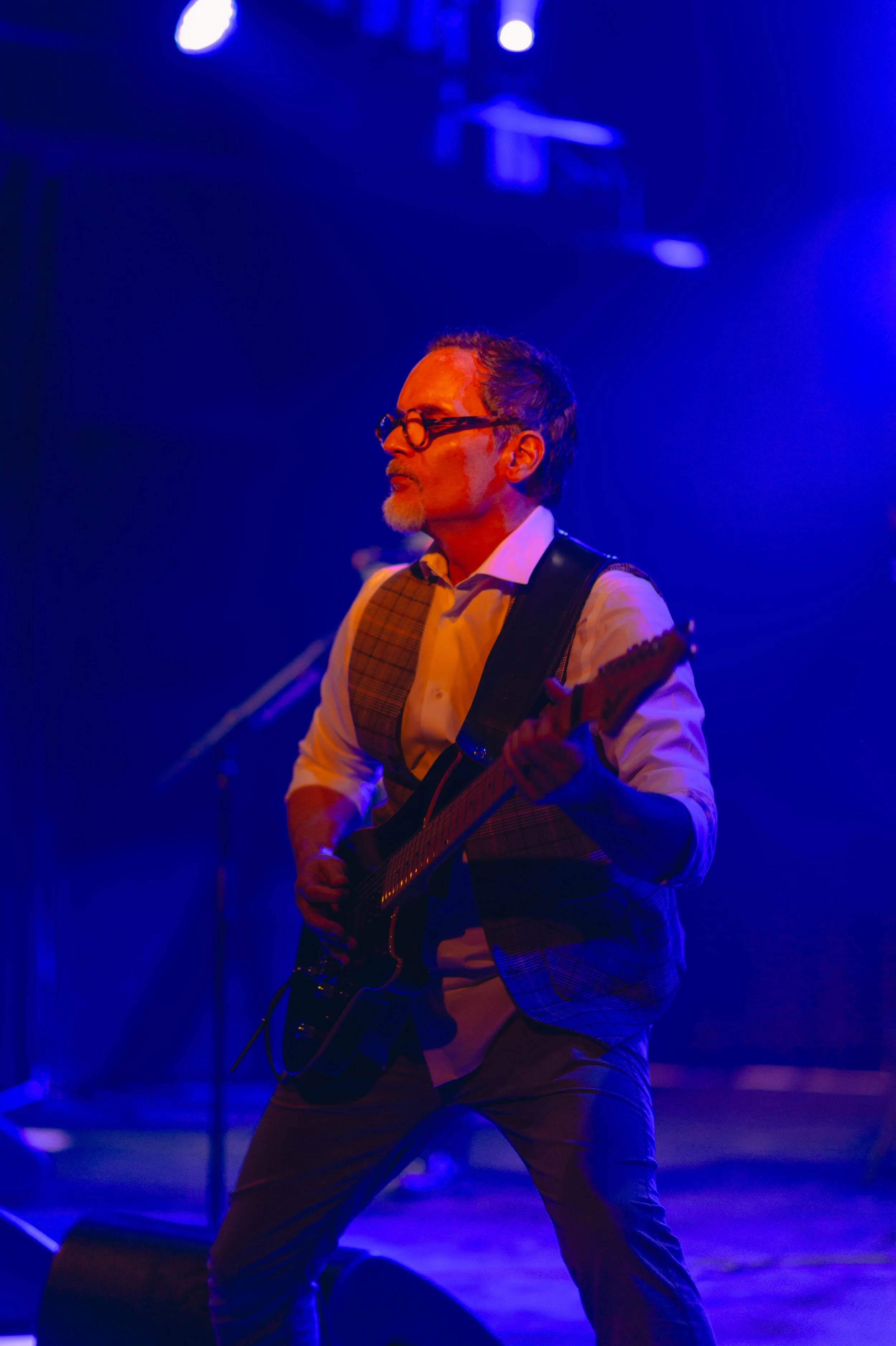 Un homme jouant de la guitare sur scène, éclairé en bleu, portant une chemise blanche, un gilet à carreaux et des lunettes, dans un environnement de concert.