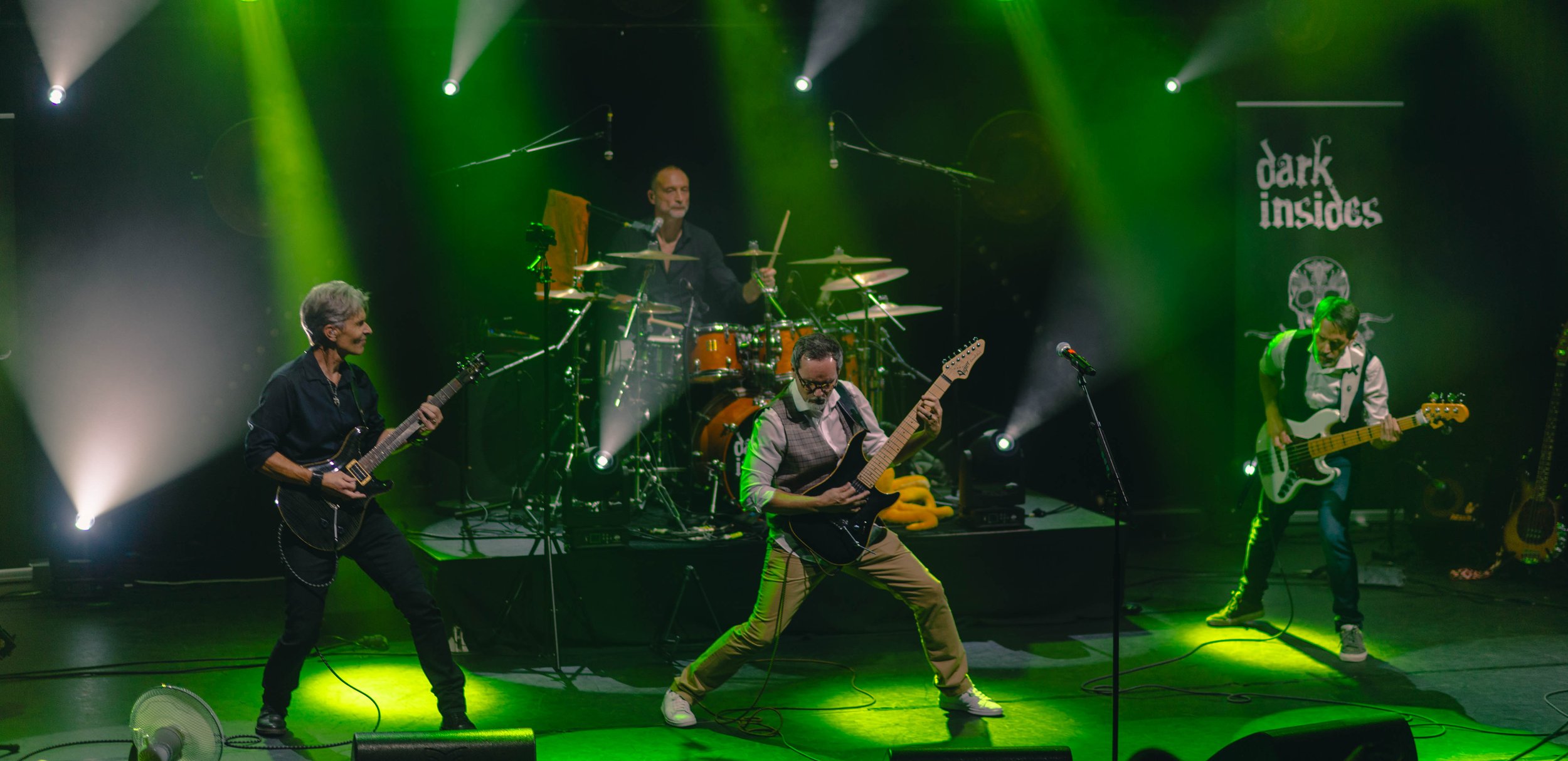 Performance de groupe musical avec quatre membres jouant de la guitare, basse et batterie sur scène avec éclairage vert et noir, logo 'Dark Inside' en arrière-plan.