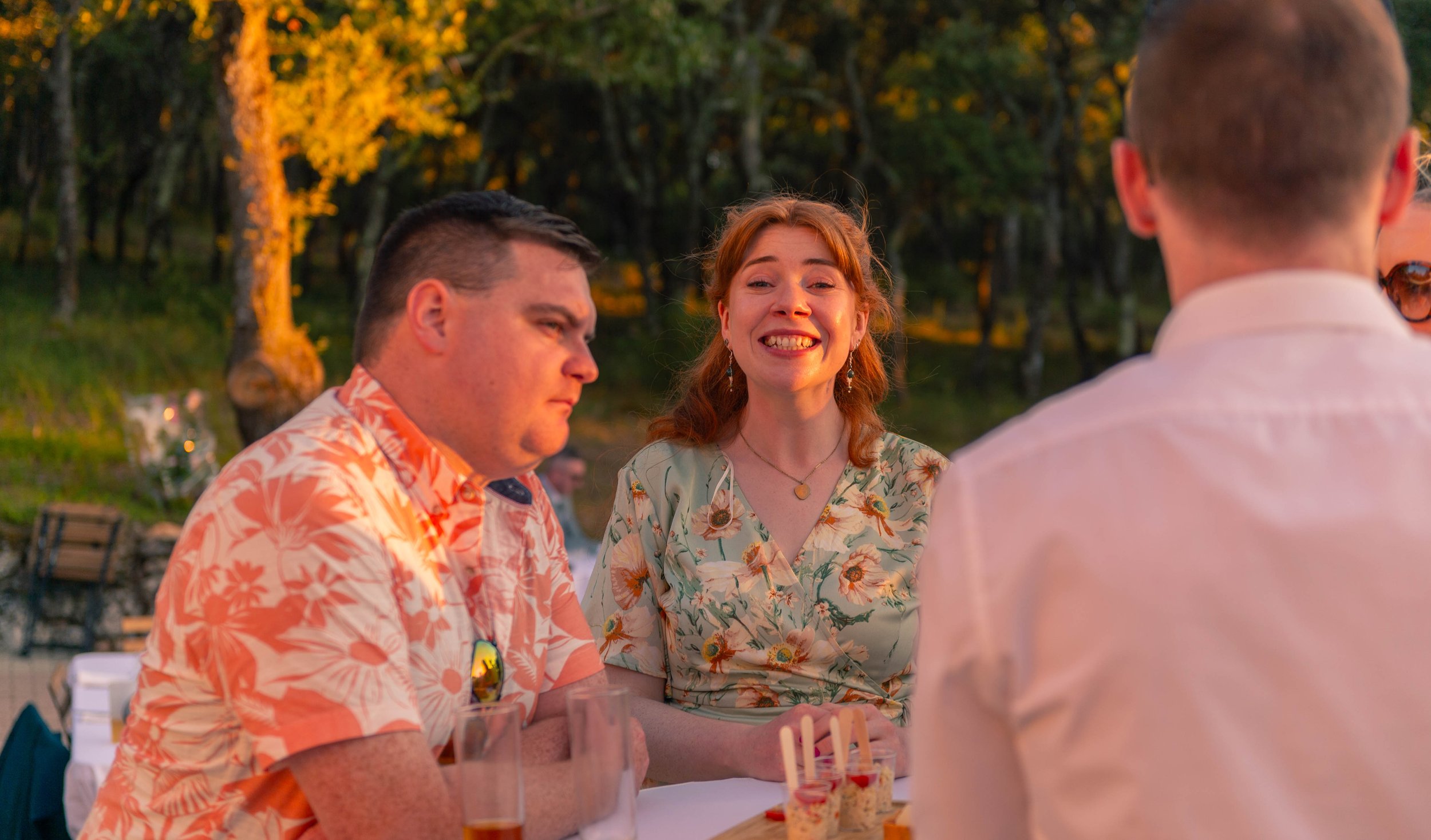 Groupe de personnes assises à une table en plein air lors d'une fête ou d'un rassemblement, avec des forêts en arrière-plan, ambiance chaleureuse et décontractée au coucher du soleil.
