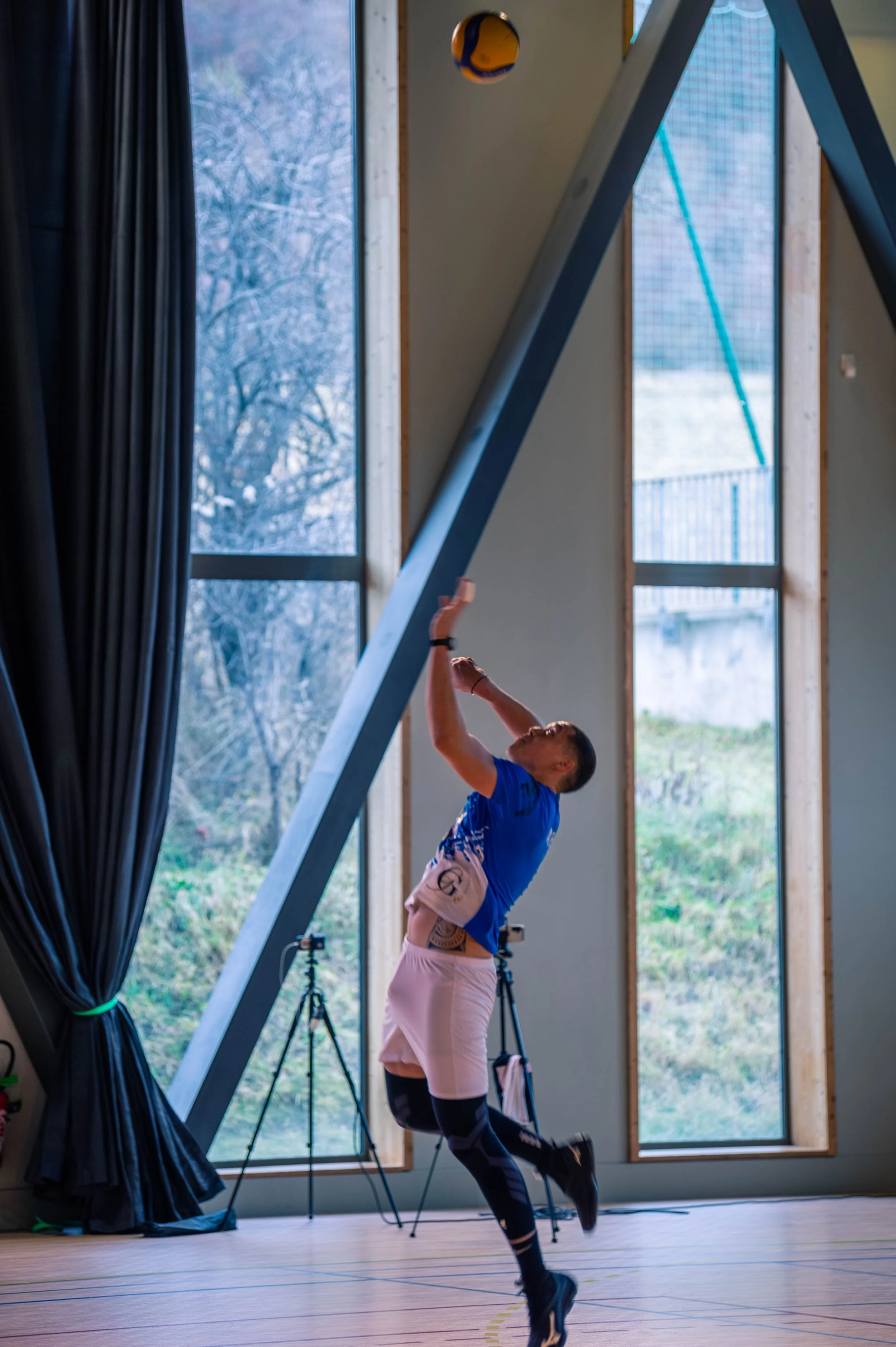 Un homme en tenue sportive joue au volley-ball en intérieur, avec de grandes fenêtres en arrière-plan et un filet de volley-ball suspendu au plafond.