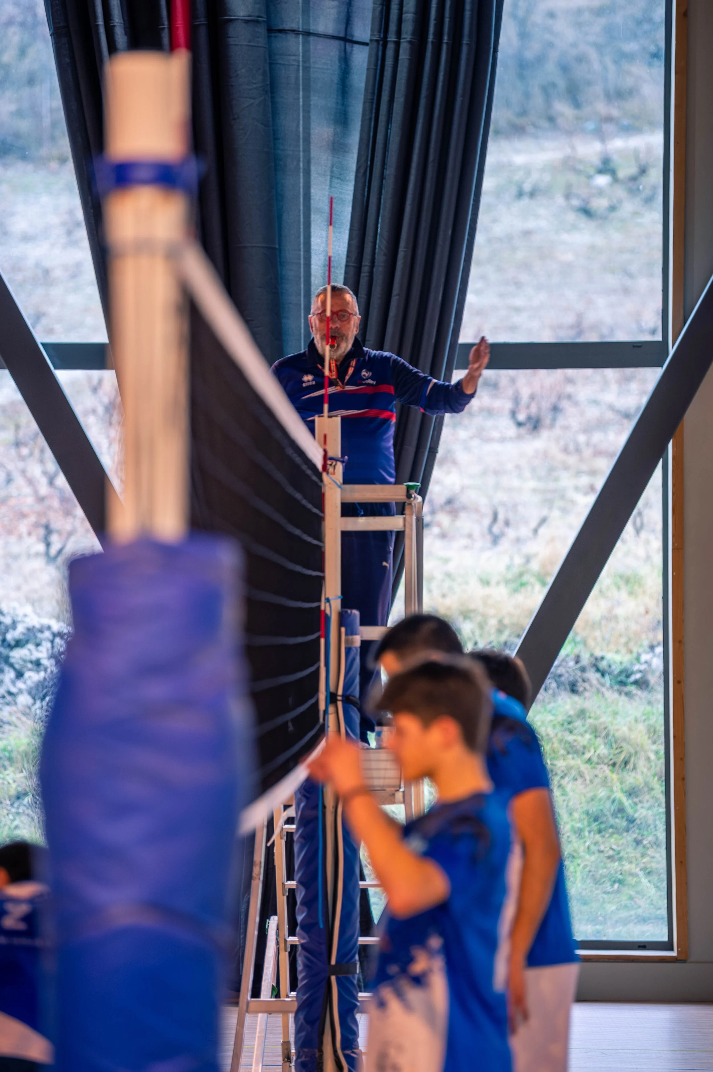 Un entraîneur ou un arbitre de volleyball déployant une échelle pour ajuster ou vérifier un filet de volleyball au sein d'une salle, avec des jeunes joueurs en uniforme en avant-plan.