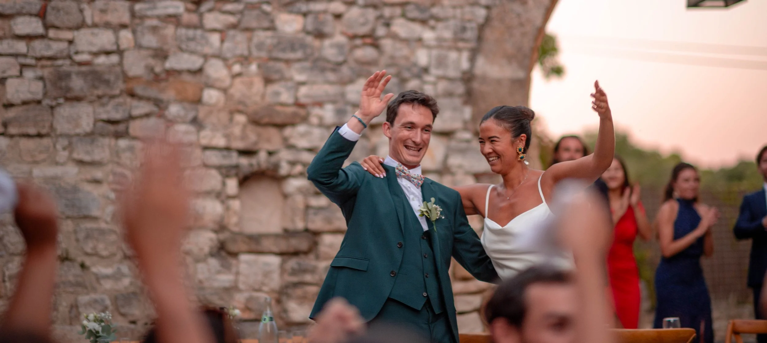 Un couple de mariés danse lors d'un mariage en plein air avec des invités en arrière-plan, contre un mur en pierre, pendant le coucher du soleil.
