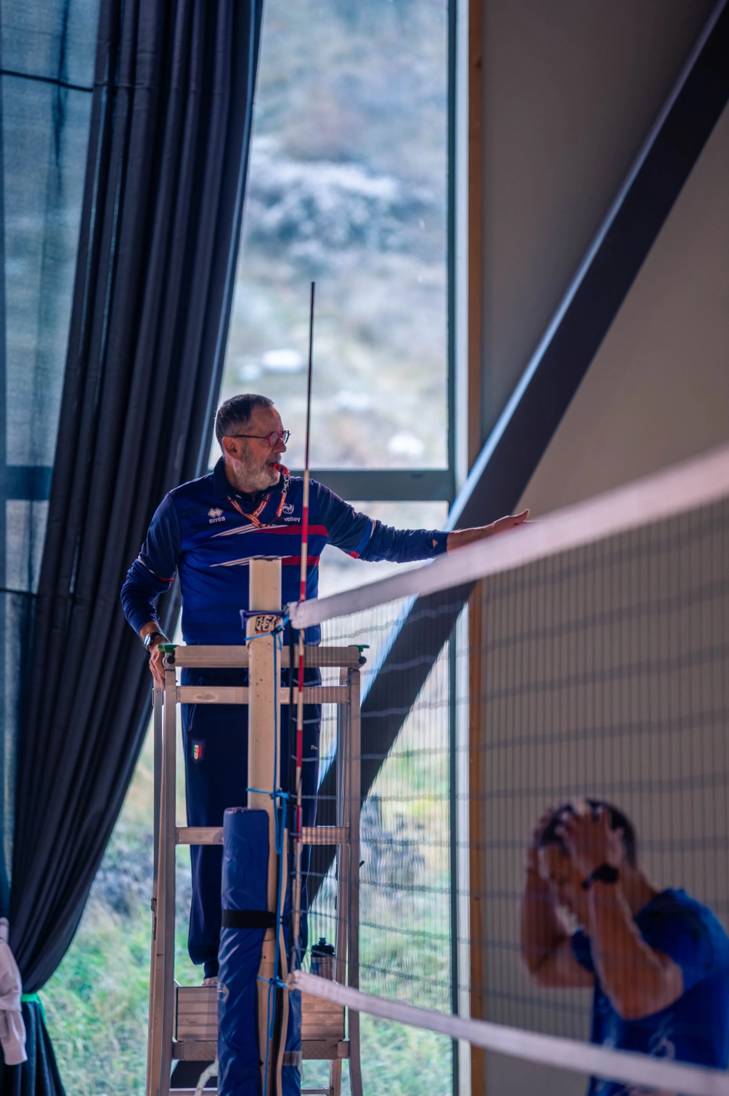 Un entraîneur supervise un match de volleyball féminin à l'intérieur, avec une joueuse en difficulté visible au premier plan.