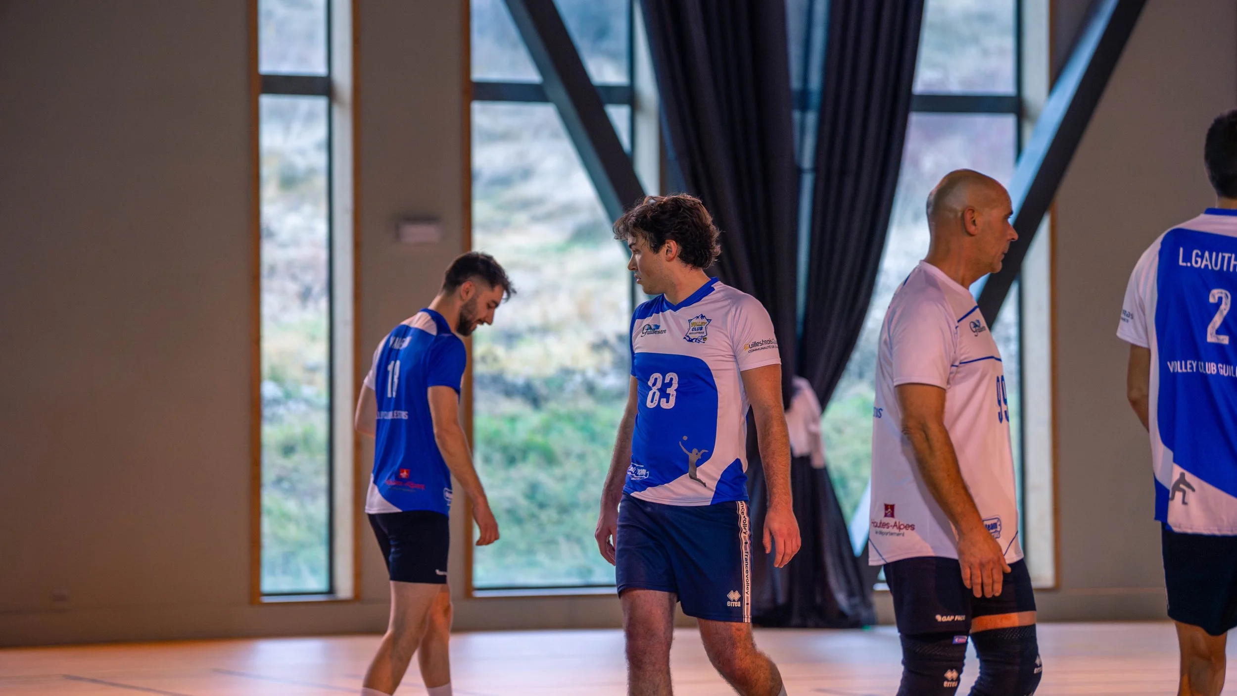 Équipe de volleyball en tenue bleue et blanche sur un terrain intérieur, certains joueurs manifestant des émotions, au gymnase avec grands fenêtres et rideaux noirs en arrière-plan.