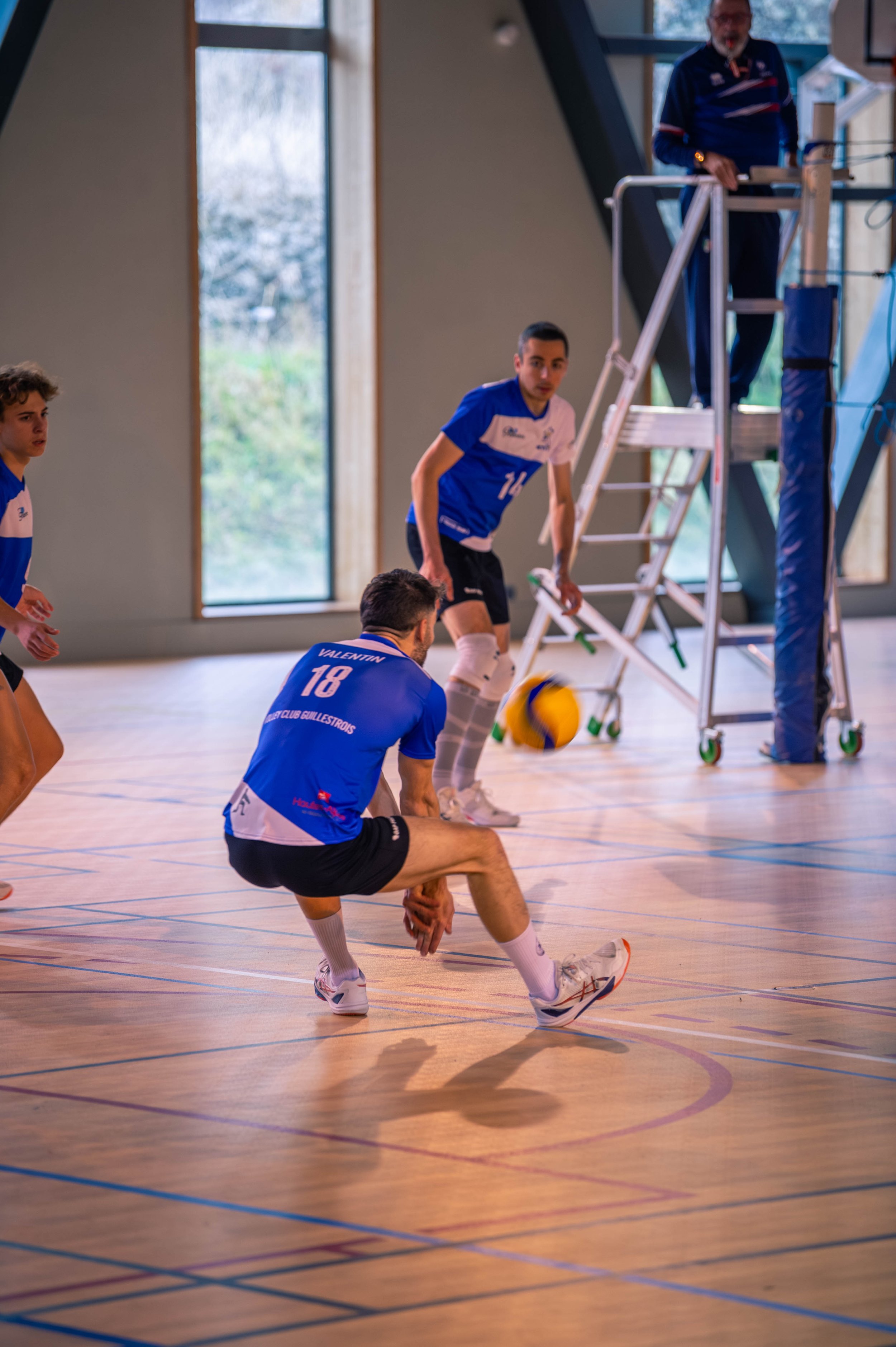 Des joueurs de volley-ball en action dans une salle, avec un ballon en vol, dont un est assis sur le sol, tandis que d'autres se préparent à jouer. Un entraîneur ou arbitre est visible en arrière-plan, près d'un escabeau.