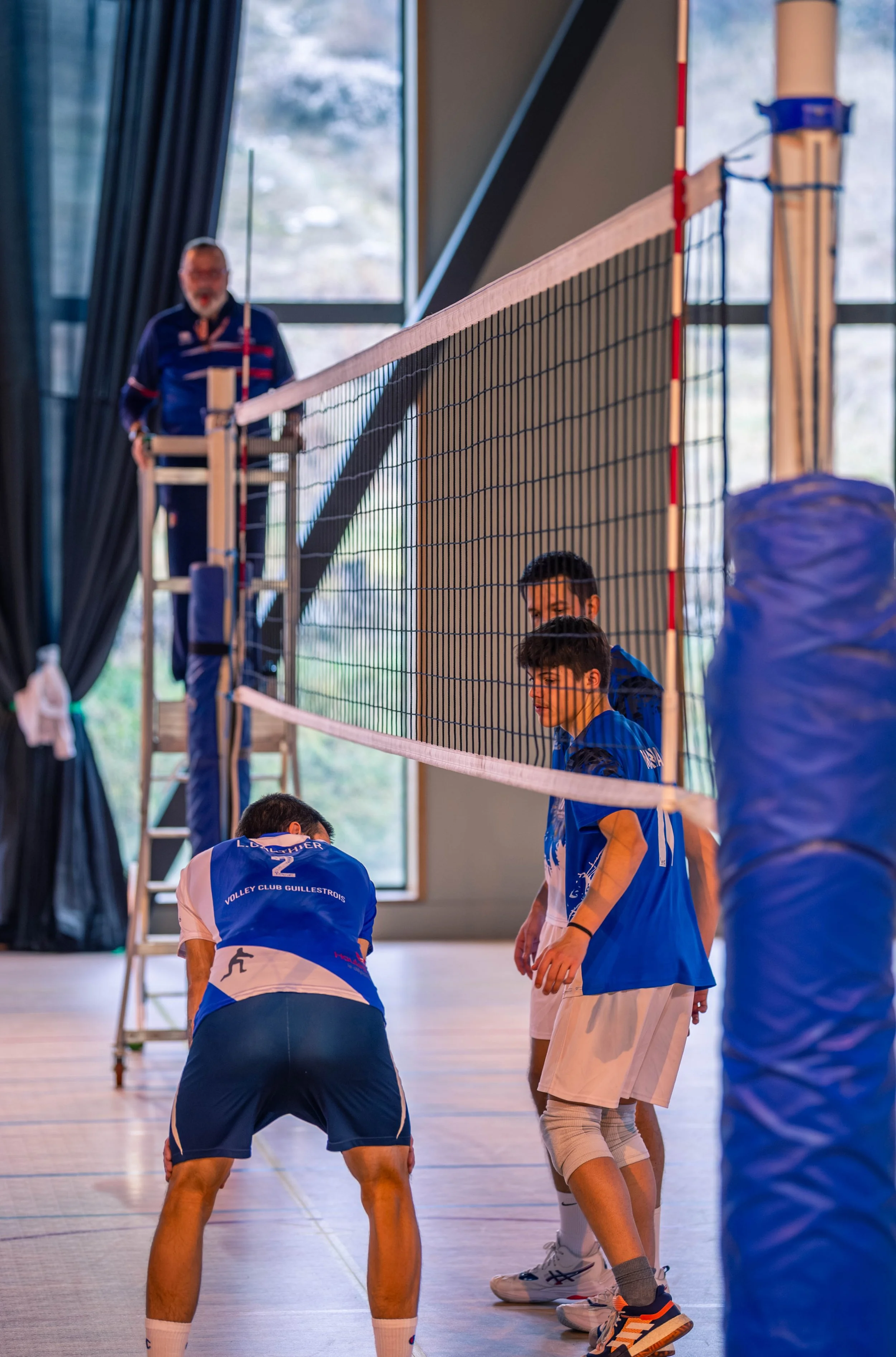 Des joueurs de volley-ball en entraînement à l'intérieur, portant des maillots bleus, près d'un filet de volleyball, avec un entraîneur en arrière-plan.