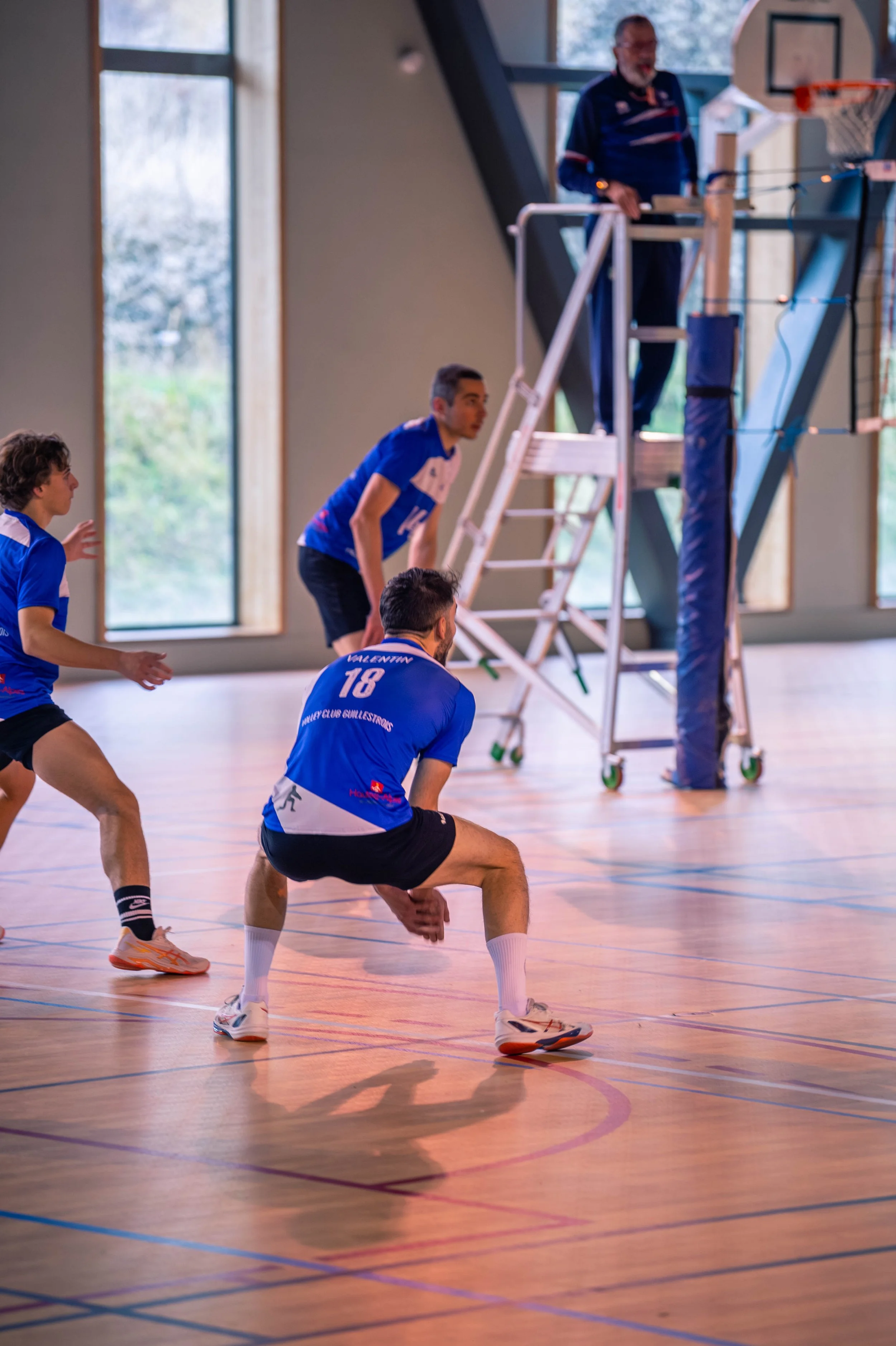 Une équipe de volleyball en pleine action à l'intérieur d'un gymnase, avec un entraîneur observant depuis une plateforme en hauteur.