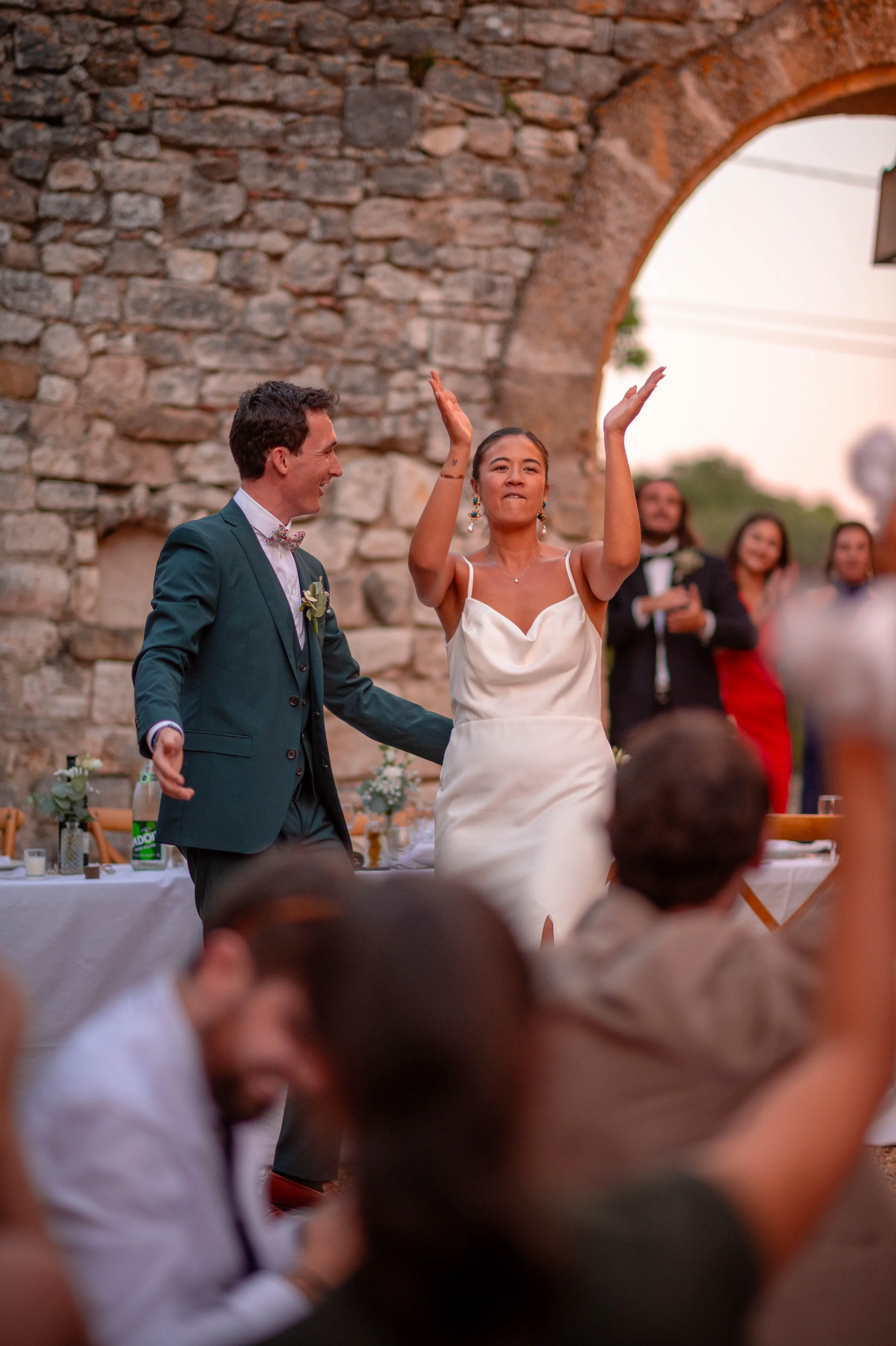 Une femme en robe de mariage et un homme en costume discutent lors d'une célébration en plein air, entourés d'invités.