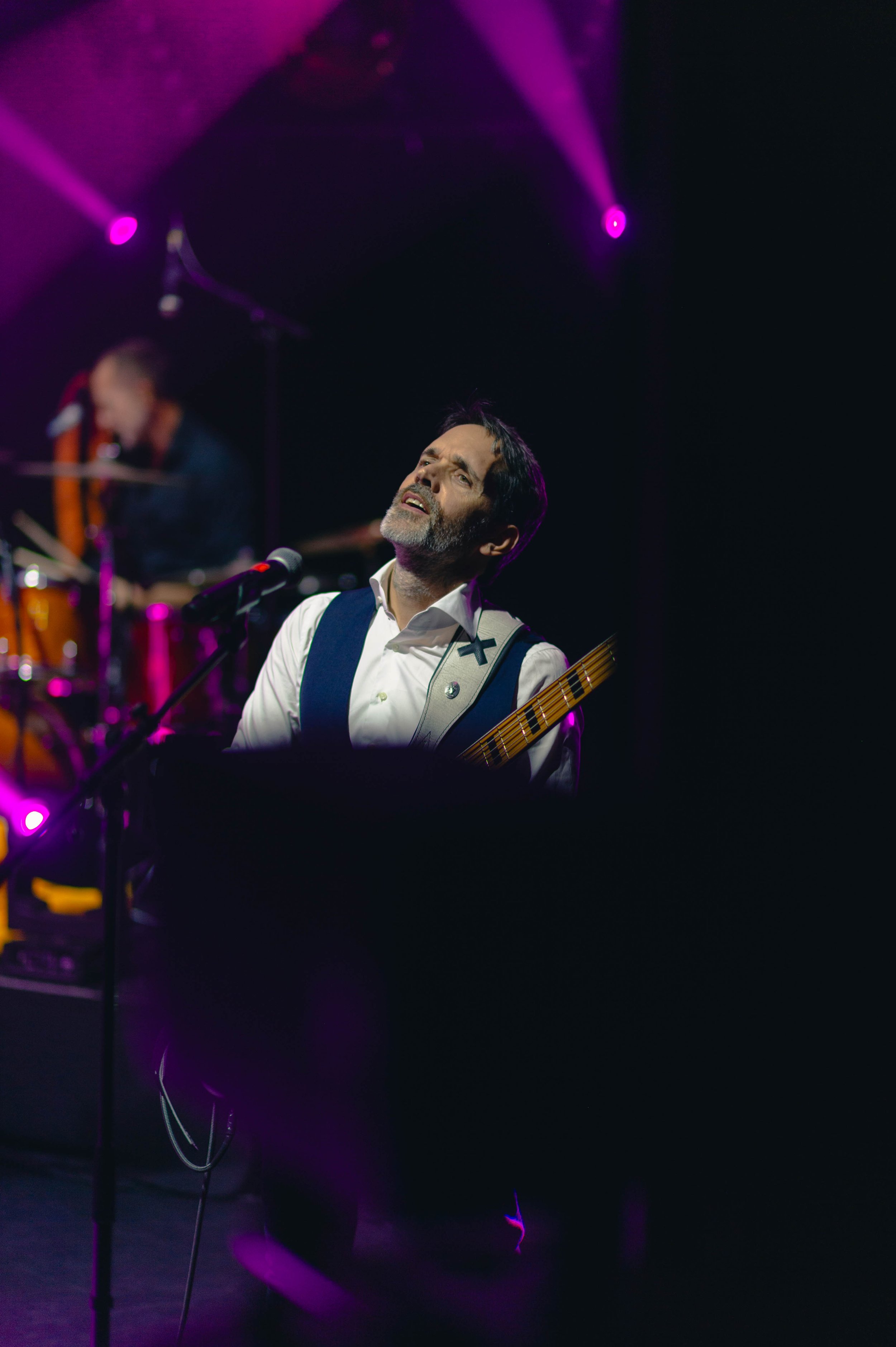 Un musicien chante lors d'un concert, avec un guitariste en arrière-plan et des lumières violettes au plafond.