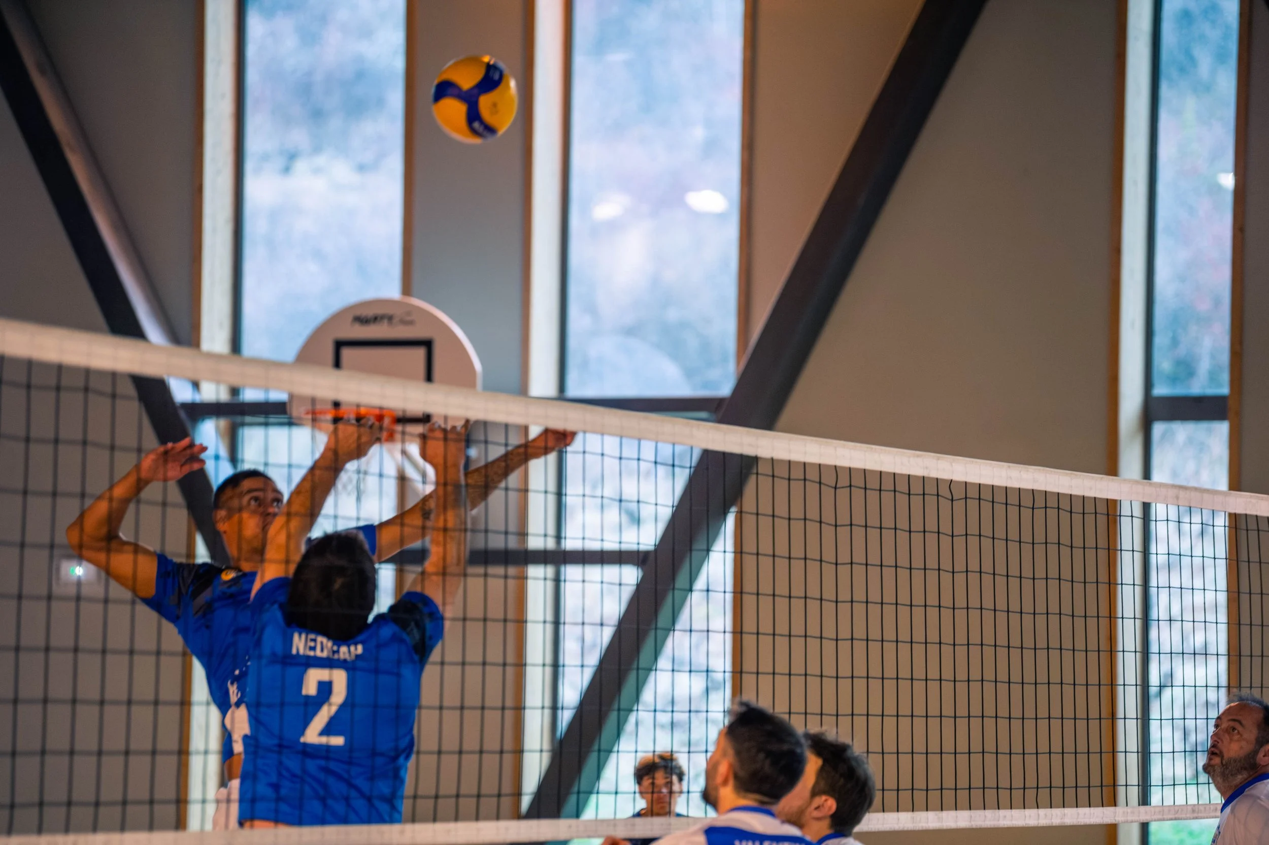 Une scène de volleyball en intérieur avec des joueurs en action, deux joueurs sont en train de sauter pour bloquer la balle au-dessus du filet, avec des spectateurs en arrière-plan.