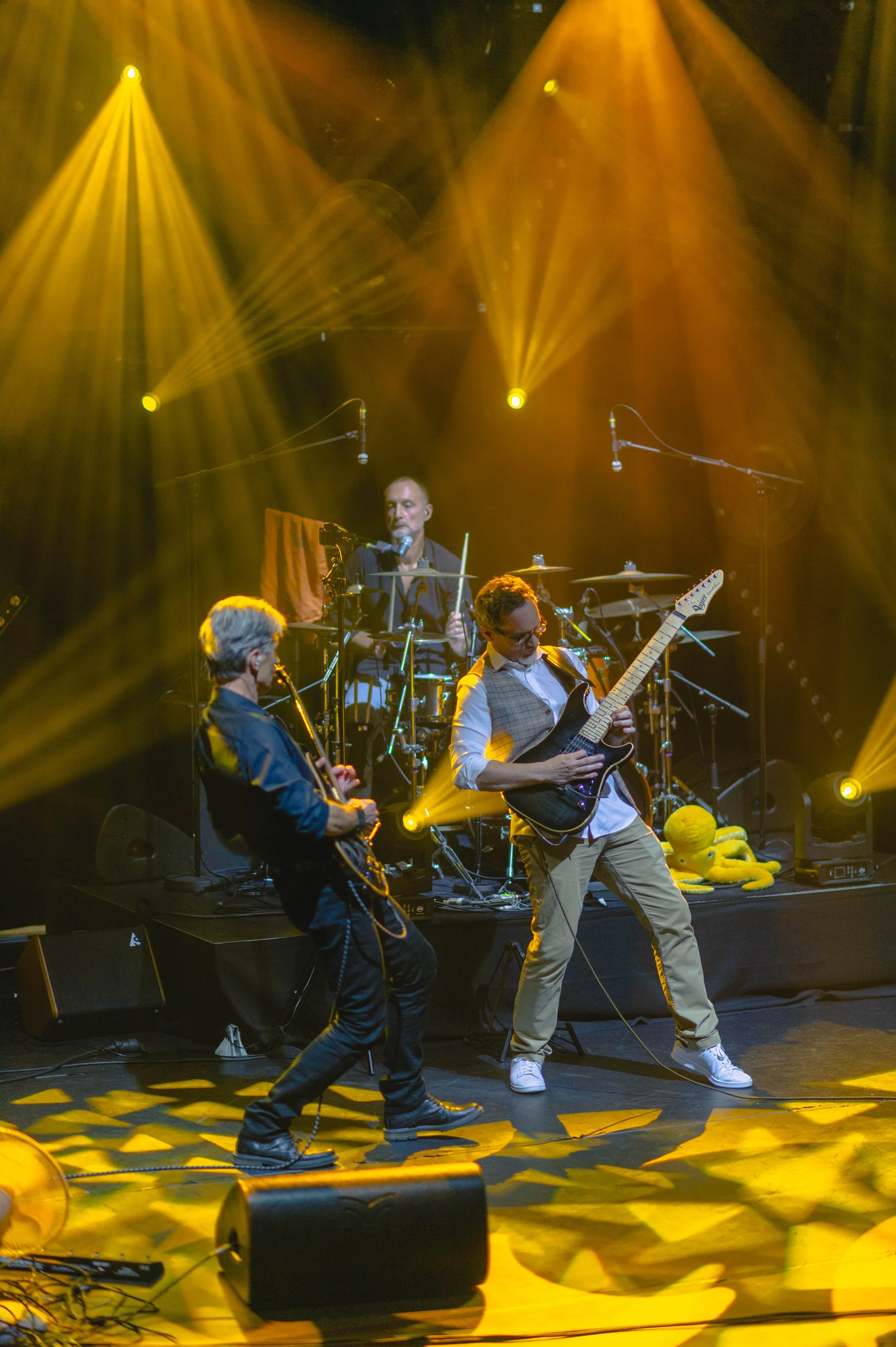 Trois musiciens jouent sur scène sous des lumières jaunes et orange, avec un batteur au fond, deux guitaristes devant, un avec une guitare électrique et l'autre avec une guitare acoustique. Il y a un jouet en peluche jaune sur la scène à droite.