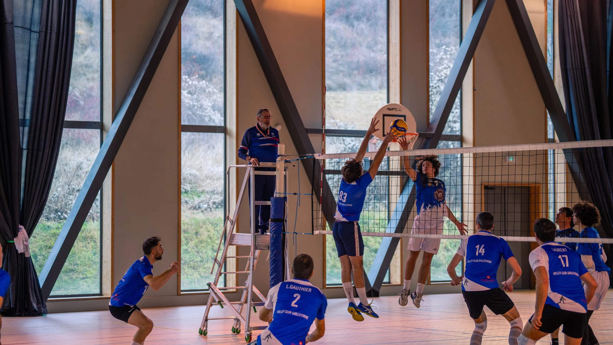 Un match de volleyball en intérieur avec des joueurs en action, certains sautant pour attaquer ou bloquer la balle, sous un éclairage naturel provenant de grandes fenêtres.