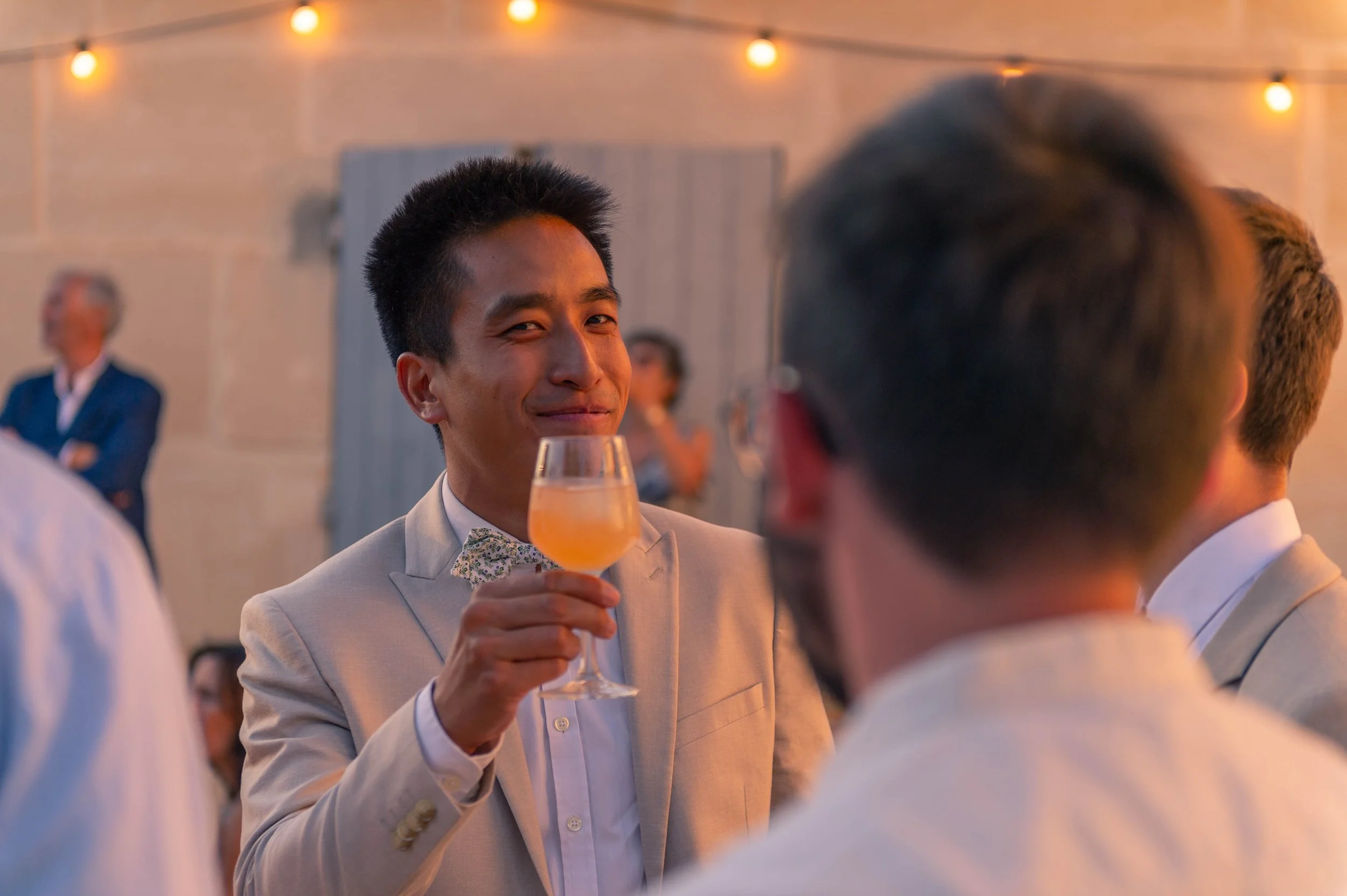 Groupe de personnes lors d'une fête, un homme souriant en costume clair tenant un verre de cocktail orange.