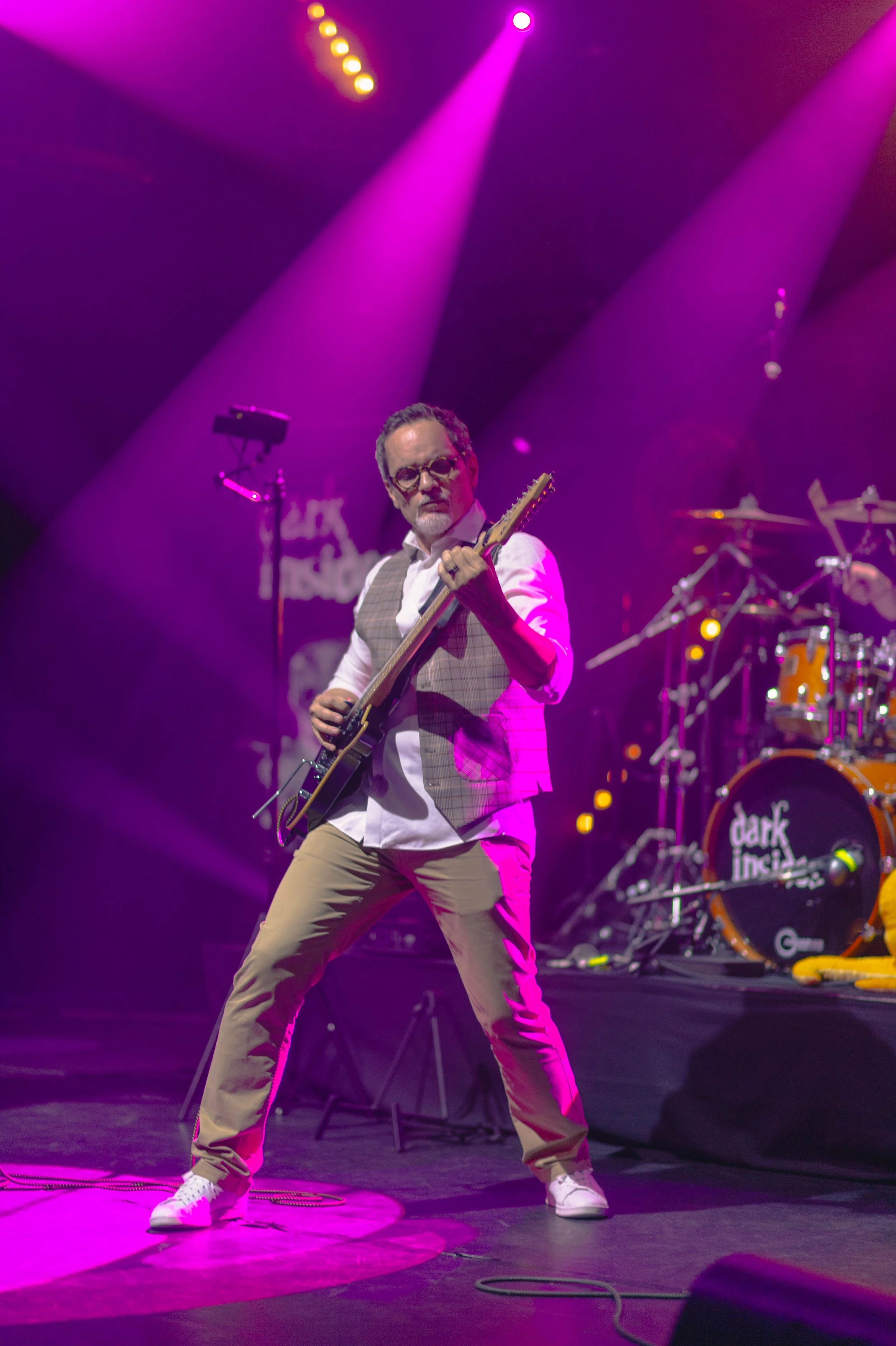 Musicien jouant de la guitare électrique sur scène avec éclairages violets et un batteur en arrière-plan, logo du groupe 'dark impulse' visible sur la batterie.