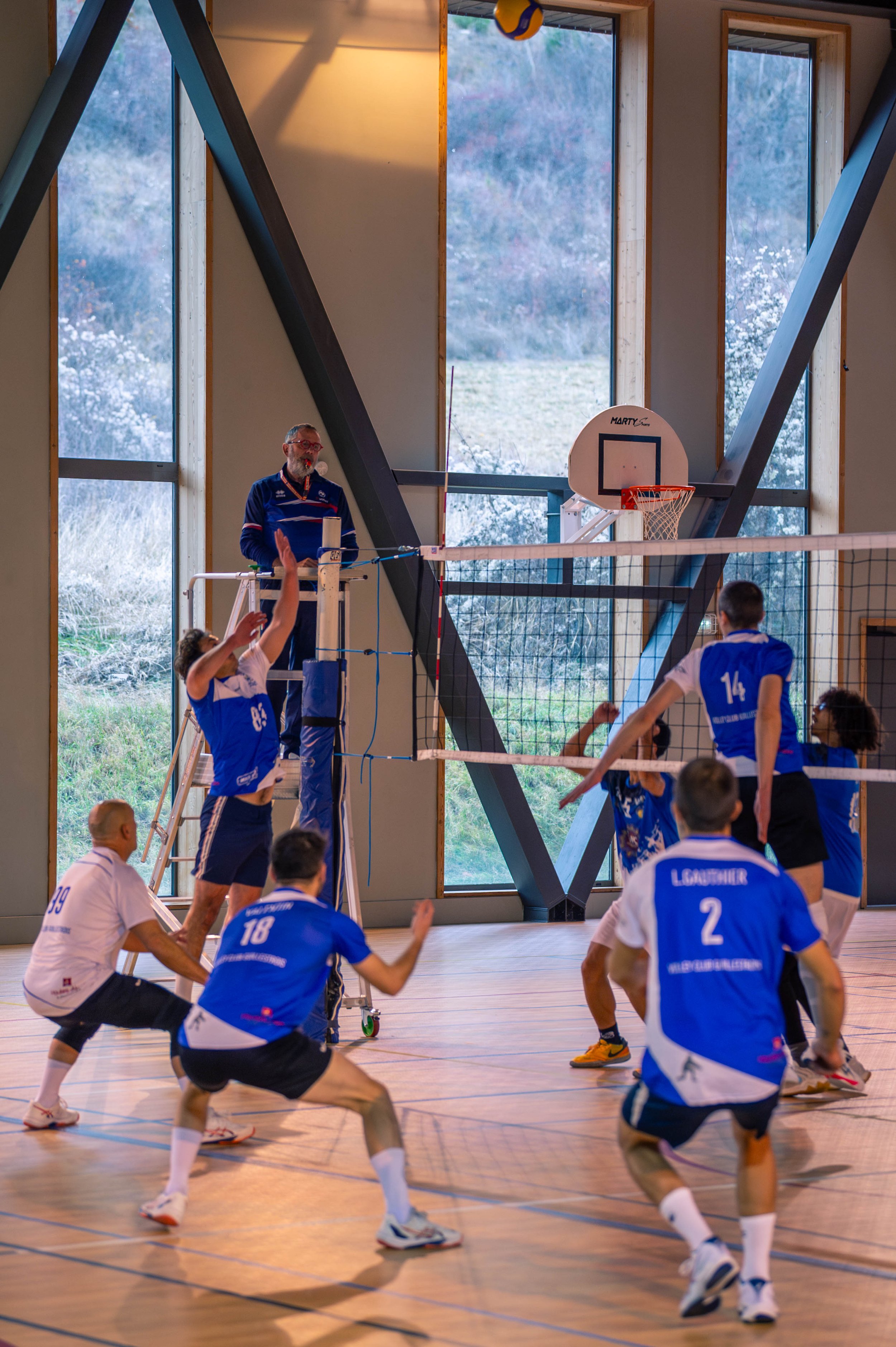 Une équipe de volleyball en train de jouer dans un gymnase intérieur avec de grandes fenêtres, un arbitre sur une plateforme, et un ballon suspendu au-dessus du filet.
