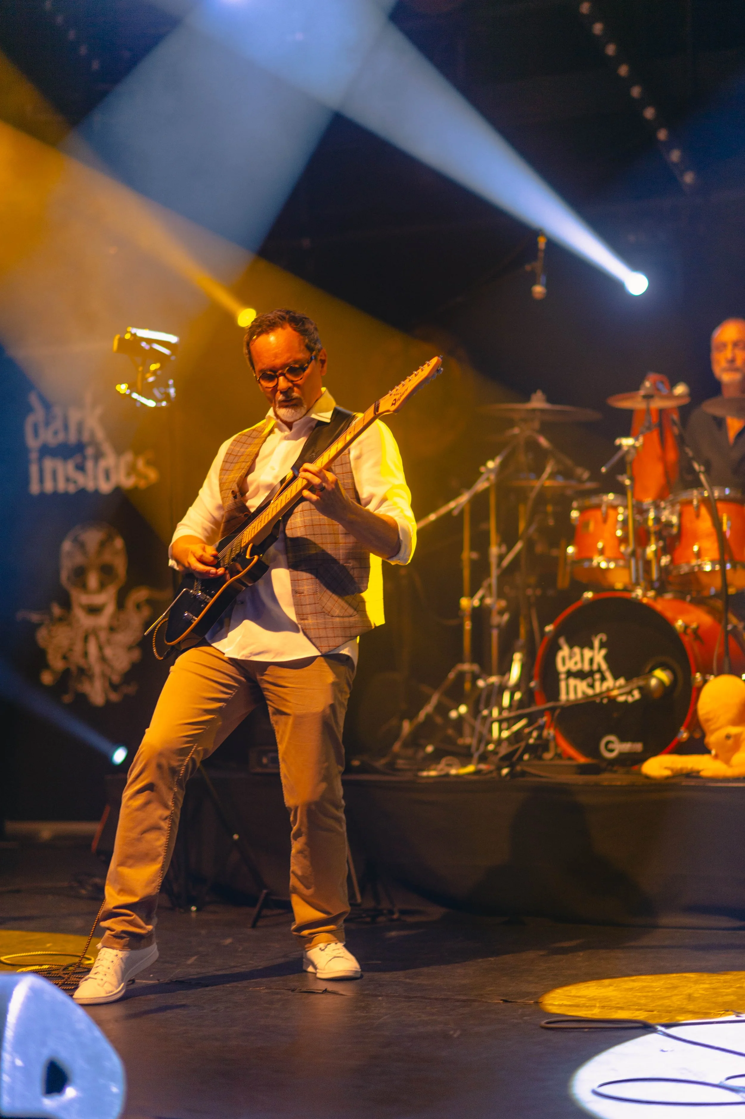 Un musicien joue de la guitare électrique sur scène avec un groupe, éclairé par des lumières colorées.